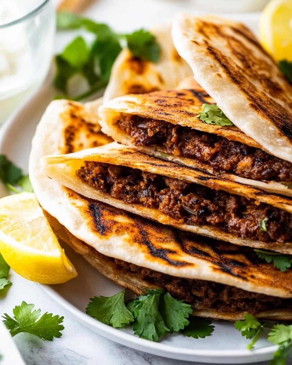 The image shows three grilled flatbreads filled with a dark brown, finely ground meat mixture, stacked close together on a white plate. Each flatbread is folded in half with the filling visible along the edges, displaying a crispy and slightly charred golden-brown exterior. Fresh green cilantro leaves are scattered around the flatbreads and on top, adding a pop of bright color. A lemon wedge is placed near the plate's edge, and part of a glass of water is visible on the left side. The background features a white marbled surface with hints of blurred green herbs and a dollop of white sauce in another white dish in the background. Photo taken with an iphone --ar 4:5 --v 6.1