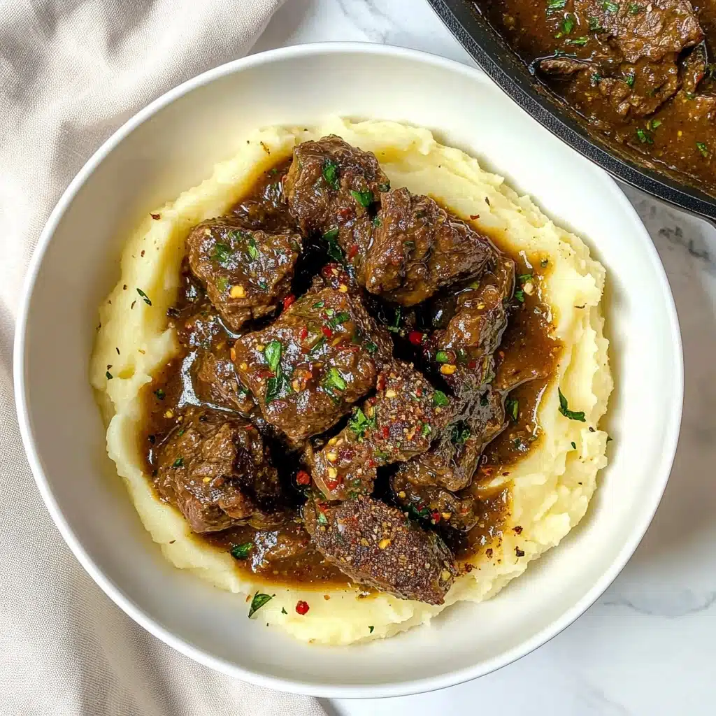 Slow Cooker Garlic Butter Steak Bites, garlic butter steak, slow cooker steak recipe, tender steak bites, easy beef dinner The image shows a white plate with a bed of creamy mashed potatoes forming the bottom layer, smooth and light in texture. On top, there is a generous pile of glossy, golden-brown beef cubes in a rich brown gravy, sprinkled with small bits of green parsley for color contrast. A fork with a blue handle rests on the edge of the plate. In the background, fresh green celery leaves and a whole onion are placed on a white marbled surface.