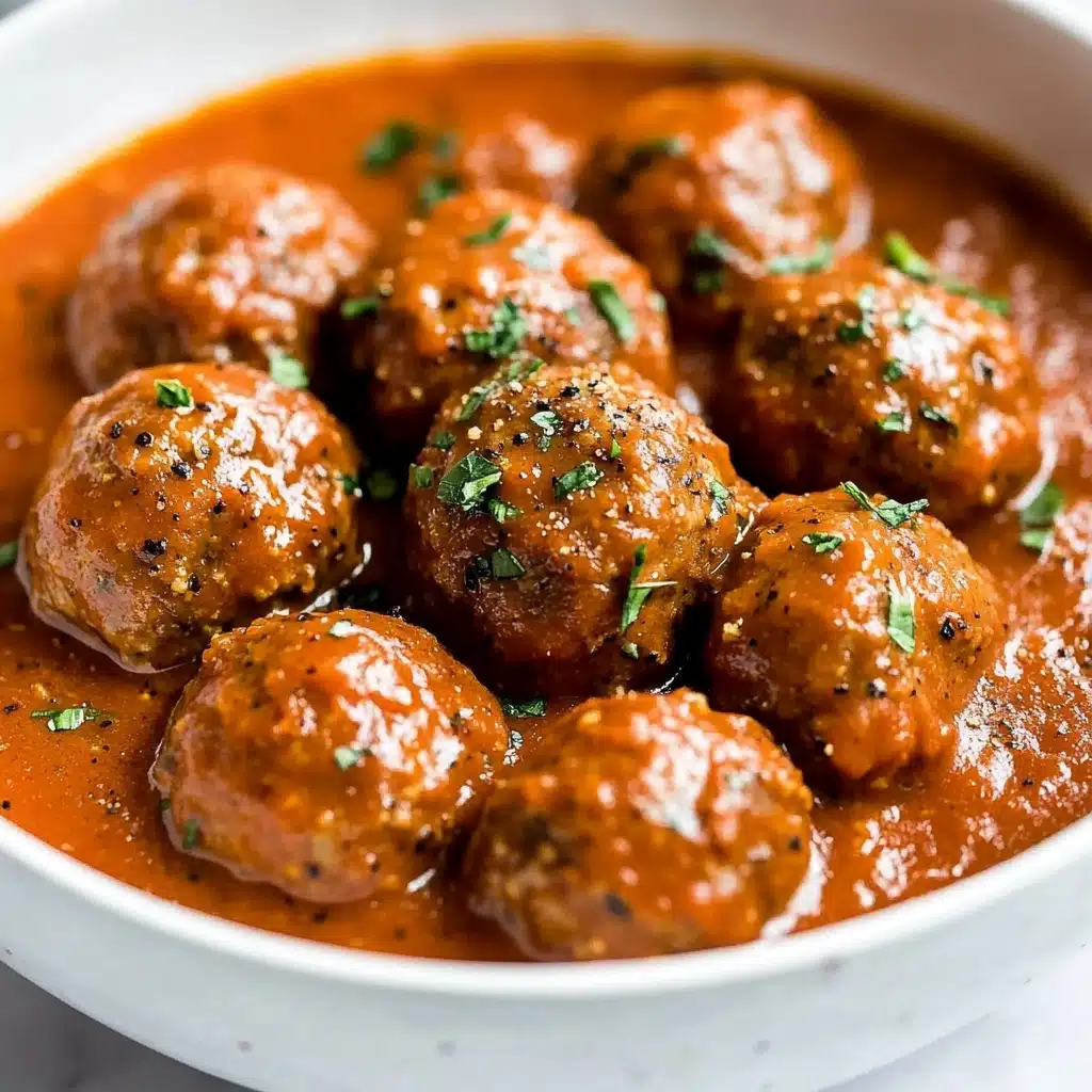Saucy Beef and Lentil Meatballs, beef and lentil meatballs, hearty meatball recipe, healthy meatball dinner, flavorful tomato coconut sauce The image shows round meatballs covered in a thick, smooth reddish-brown sauce inside a white bowl. The meatballs have a slightly shiny, moist texture, and the sauce looks creamy with a few small black pepper specks and green herb bits scattered on top. The meatballs are closely packed in the bowl, with the sauce filling the spaces between them. The bowl sits on a white marbled surface.