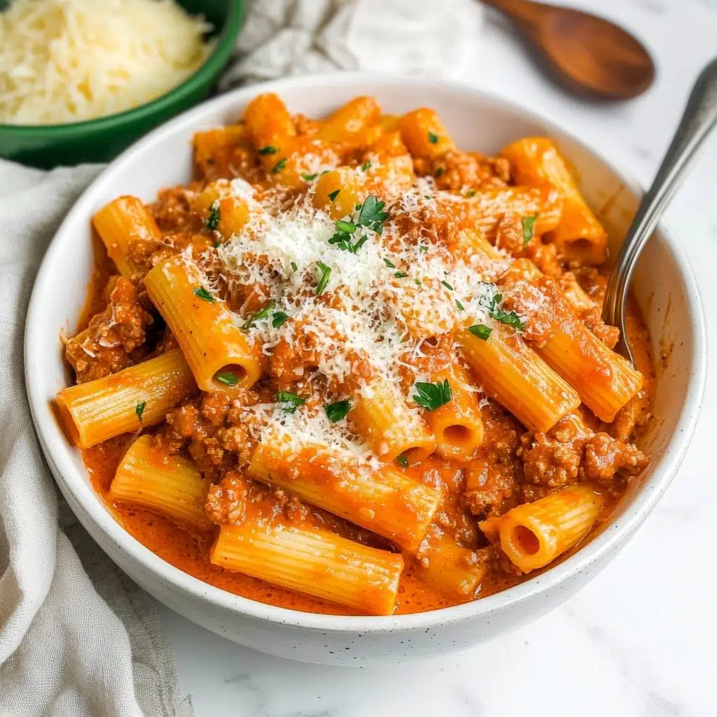 Date Me Creamy Sausage Rigatoni, creamy sausage pasta, rigatoni pasta recipes, comforting Italian pasta dishes, easy sausage pasta A white bowl filled with creamy orange-red sauce pasta, showing rigatoni pasta tubes covered in a thick sauce mixed with browned ground meat pieces. On top, a light sprinkle of grated white cheese and small green herb leaves adds color contrast. A spoon with a long silver handle is partially inside the bowl on the right side. Behind the bowl, there is a small green dish holding more grated cheese and a wooden spoon with dark brown grain resting next to it. The bowl and items are placed on a white marbled surface with a pale cloth on the left side.