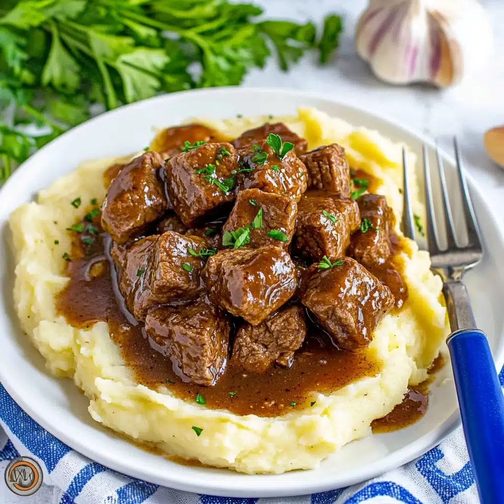 Slow Cooker Garlic Butter Steak Bites, garlic butter steak, slow cooker steak recipe, tender steak bites, easy beef dinner A white plate shows a creamy pale yellow base layer of smooth mashed potatoes spread evenly, topped with several pieces of dark brown cooked meat with a shiny, slightly oily surface. The meat is covered by a rich dark sauce with visible small bits, sprinkled with finely chopped green herbs and tiny red chili flakes, adding color contrast. Next to the plate, a black pan holds more pieces of meat in the same dark sauce. The setup is on a white marbled surface with a light beige cloth nearby.