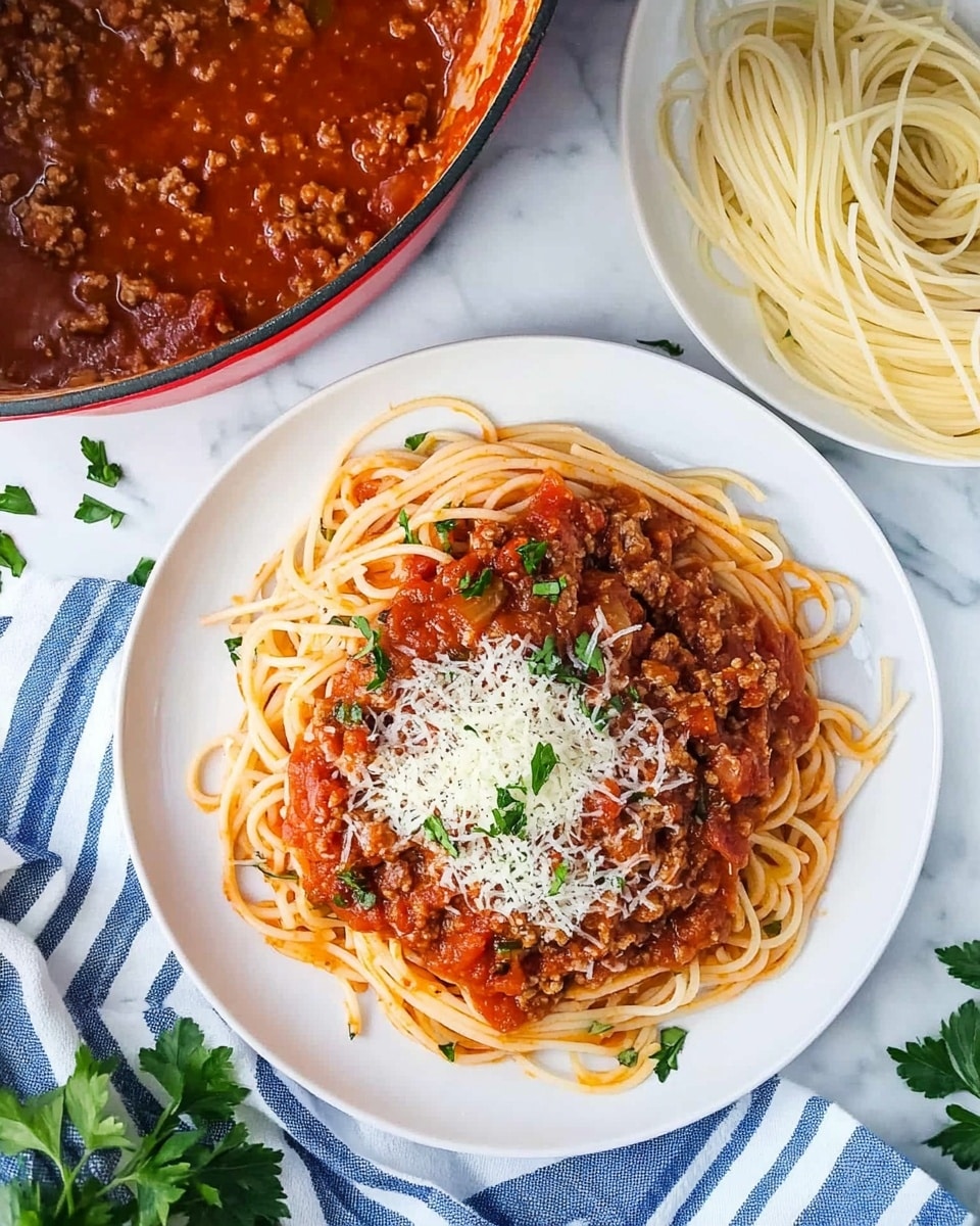 Turkey Spaghetti with Tomato Sauce and Parmesan, healthy turkey pasta, quick weeknight dinner ideas, savory turkey pasta recipes, easy turkey spaghetti A white plate holds a dish with three main layers: a bottom bed of light brown cooked spaghetti noodles, a thick and chunky bright red tomato meat sauce covering the noodles on top, and a sprinkling of grated pale yellow cheese along with chopped green herbs scattered over the sauce. The plate sits on a wooden surface with some green herb leaves around, and a striped cloth with dark blue and white colors is partly visible in the background on one side. A shiny silver pot with more spaghetti inside is placed in the background on the right side.