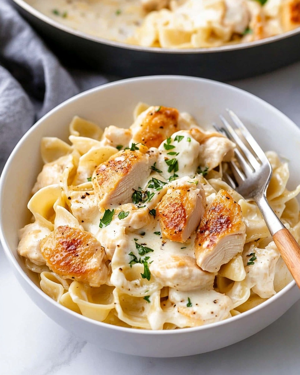 Chicken Tortellini Alfredo, creamy tortellini chicken meal, easy Alfredo pasta with chicken, homemade chicken Alfredo sauce, comforting chicken tortellini dish The image shows a close-up of a dish with about two layers: the first layer is soft, folded pasta with a creamy white sauce that looks smooth and thick, creating a rich base. On top, scattered unevenly, are thick pieces of cooked chicken, golden-brown on the outside with a juicy, white inside. Small green bits of parsley are spread across the chicken and pasta, adding a fresh pop of color. The whole dish sits on a white plate over a white marbled surface, giving a clean and bright look.