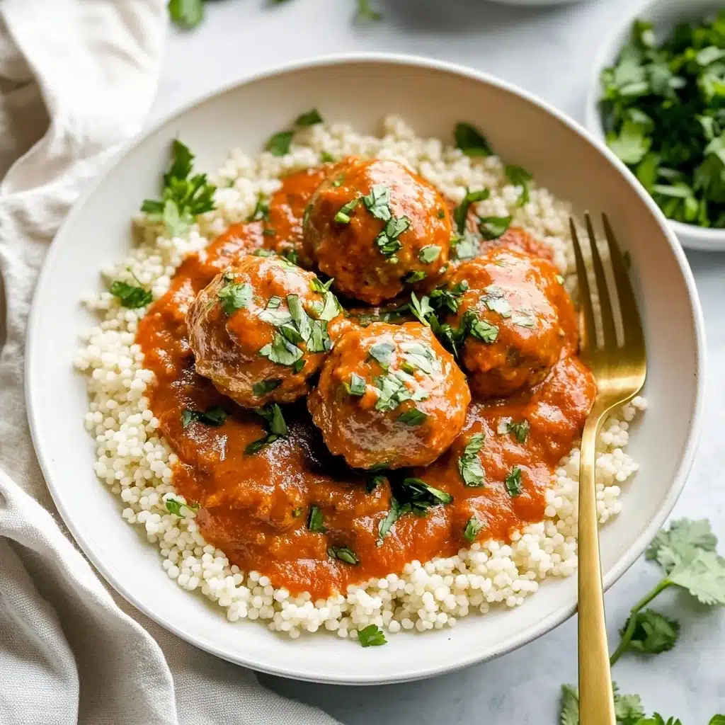 Saucy Beef and Lentil Meatballs, beef and lentil meatballs, hearty meatball recipe, healthy meatball dinner, flavorful tomato coconut sauce A white bowl filled with small round white couscous at the bottom layer, topped with five medium-sized brown meatballs covered in a smooth, thick orange-red sauce. The sauce has fresh green cilantro leaves sprinkled on top and scattered around the couscous. A gold fork rests inside the bowl on the right side. The bowl is placed on a white marbled surface with a light-colored cloth nearby.