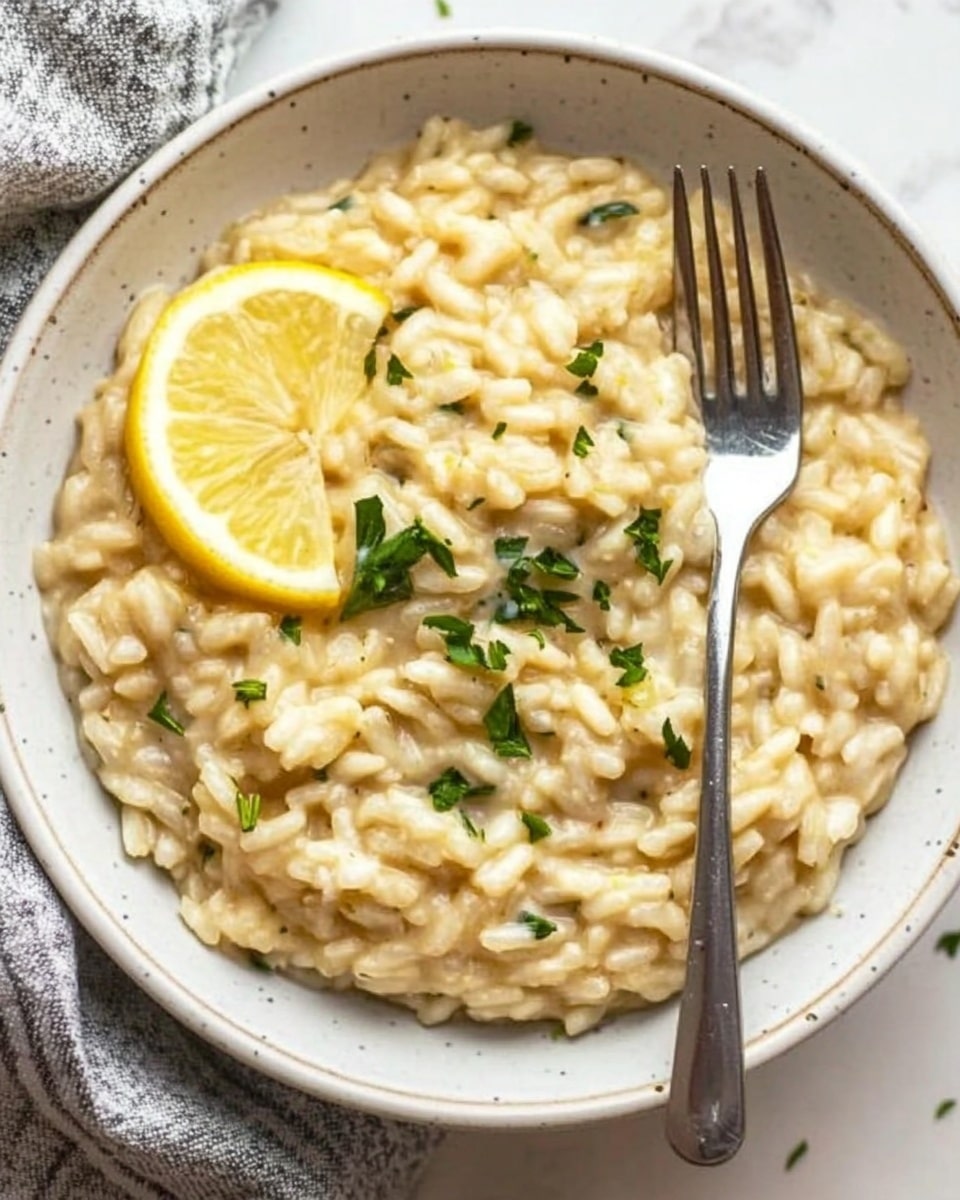 One Pot Lemon Orzo Pasta, Lemon Orzo Pasta, Easy Lemon Orzo, One Pot Pasta Recipes, Quick Lemon Pasta Dishes A white bowl filled with creamy risotto, creamy light yellow in color with small green herb pieces mixed throughout. On the left side inside the bowl, two thin lemon slices rest against the risotto. A silver fork is placed inside the bowl on the right side, partially submerged in the risotto. The bowl sits on a white marbled surface with a grey and white cloth napkin partially visible at the bottom left corner.