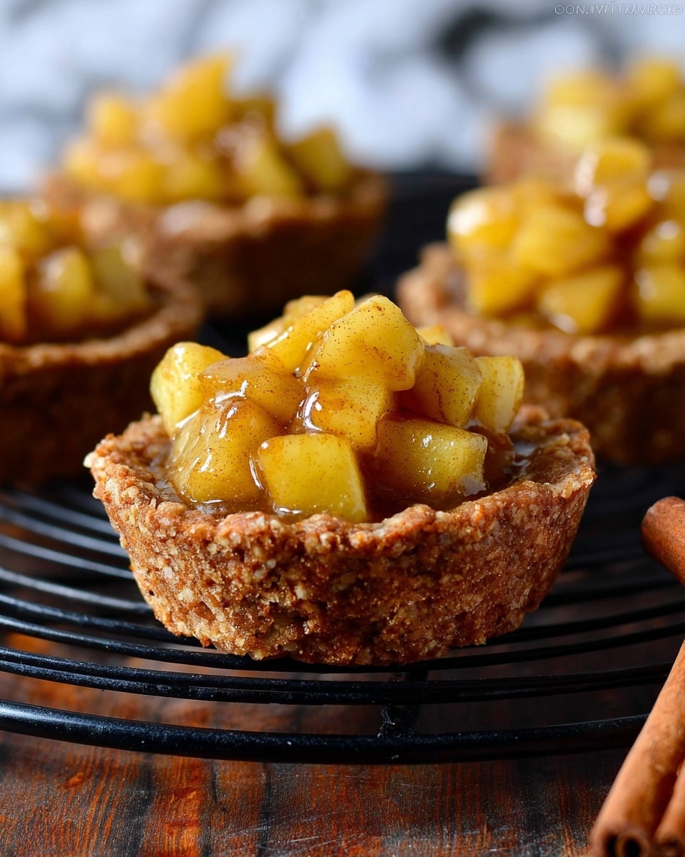 The image shows a close-up of a small tart with two layers: a rough, crumbly brown crust shaped like a cup at the bottom, and shiny, chunky yellow apple pieces coated in a glossy cinnamon sauce piled inside the crust. The tart is on a dark wooden surface with a cinnamon stick nearby, and other similar tarts are blurred in the background on a black cooling rack. The background has a white marbled texture. photo taken with an iphone --ar 4:5 --v 7