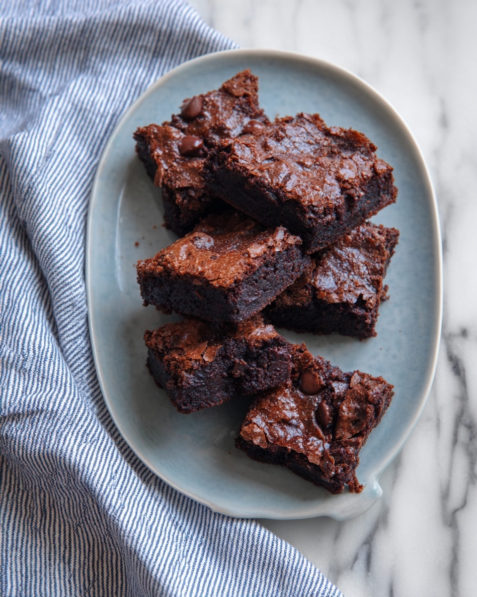 A white oval plate holds seven square-shaped dark brown brownies stacked casually. The brownies have a textured, slightly cracked surface with some shine and visible chocolate chunks inside. The plate rests on a white marbled surface with a white and blue striped cloth partially under it on the left side. The cloth is soft and slightly wrinkled, adding a cozy touch. photo taken with an iphone --ar 4:5 --v 7