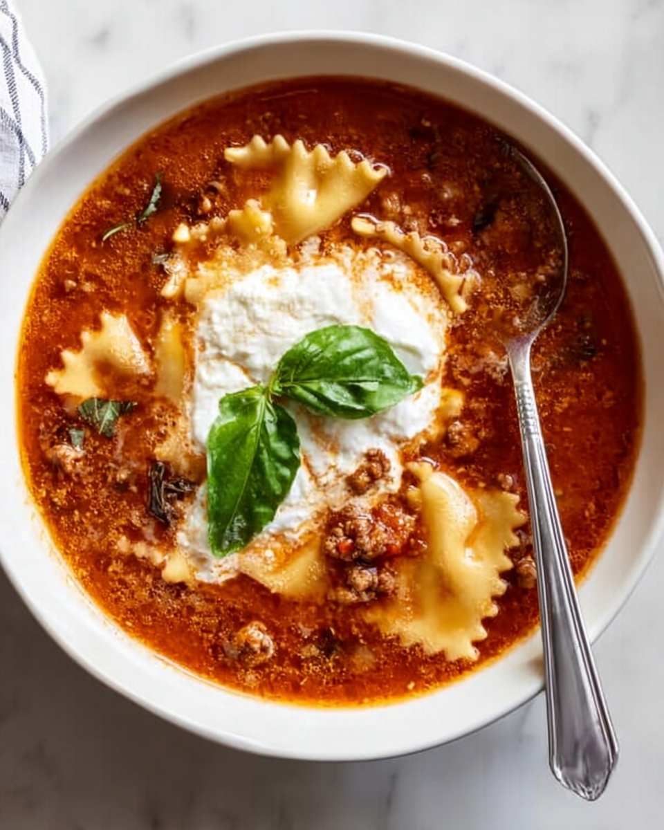 A white bowl filled with a rich, reddish-brown soup that has visible small pieces of meat and vegetables, along with wide, flat lasagna noodles partly submerged and curled at the edges. A dollop of creamy white ricotta cheese sits in the center, topped with two fresh green basil leaves. A silver spoon rests inside the bowl on the left side. The bowl is placed on a white marbled surface. Photo taken with an iphone --ar 4:5 --v 7