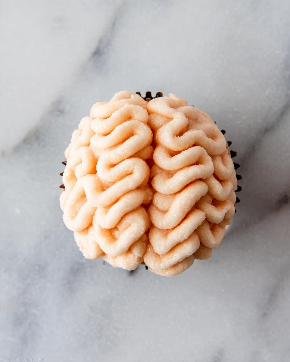 A single cupcake sits on a white marbled surface, topped with light peach icing piped in thick, wavy lines forming two parallel rows with a straight line of icing in the center, resembling a brain shape. The cupcake liner is dark-colored, providing contrast to the soft, creamy texture of the frosting, and the cupcake is viewed from above, emphasizing the detailed pattern of the frosting. Photo taken with an iphone --ar 4:5 --v 7