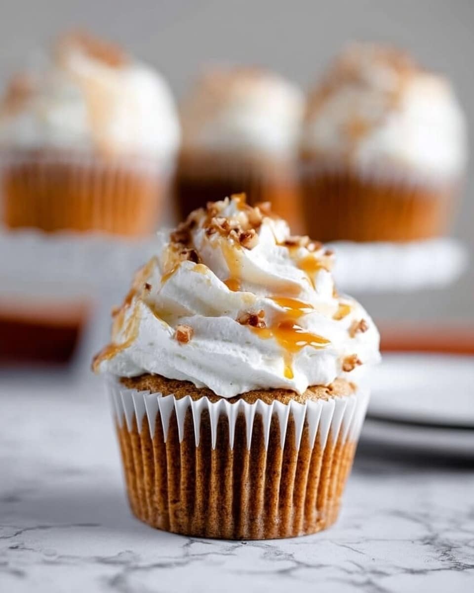 The image shows a close-up of a cupcake in a white cupcake liner on a white marbled surface. The cupcake has one layer of light brown cake at the bottom with a smooth texture. On top, there is a thick swirl of soft white frosting with a slightly fluffy look, drizzled lightly with golden caramel sauce. In the background, blurred cupcakes with similar frosting and caramel can be seen. Photo taken with an iphone --ar 4:5 --v 7