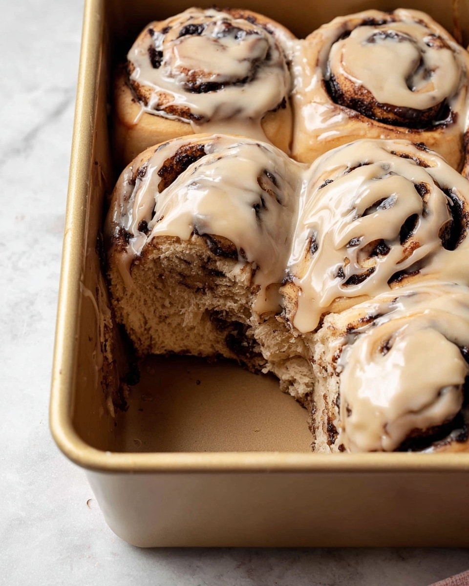 The image shows a close-up of six cinnamon rolls inside a light brown baking pan with an empty space where one roll is taken out, revealing the soft, brown interior. Each roll has dark brown swirls of cinnamon and sugar, topped generously with a light tan creamy icing that drips slightly down the sides. The rolls are packed closely together, with a soft, fluffy texture visible on the inside and a slightly shiny glaze on the icing. The background surface is a white marbled texture. Photo taken with an iphone --ar 4:5 --v 7