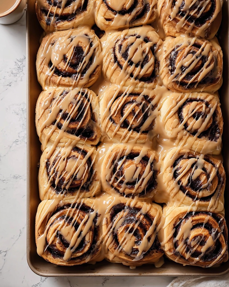 A tray filled with twelve cinnamon rolls arranged in a 3 by 4 grid, each roll showing spirals of dark brown cinnamon filling surrounded by golden-brown dough layers. A light brown glaze is drizzled over the top in thin, uneven lines, covering almost the whole surface of each roll and pooling slightly around the edges where the rolls meet. The tray sits on a white marbled surface with part of a cup visible in the top left corner. The image shows the cinnamon rolls close-up with a warm, inviting tone. Photo taken with an iphone --ar 4:5 --v 7
