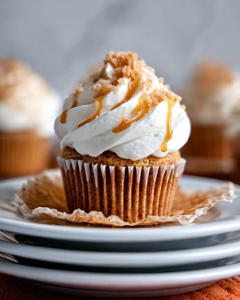 A close-up view of a single cupcake with a light brown cake base and a generous swirl of white cream frosting on top. The frosting is decorated with thin, light brown drizzle and small crunchy pieces. The cupcake sits on the slightly crumpled transparent wrapper, placed on two stacked white plates. The background is a white marbled texture with a blurred presence of other similar cupcakes behind. Photo taken with an iphone --ar 4:5 --v 7