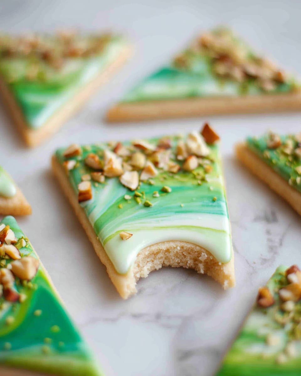 The image shows several triangular cookies spread out on a white marbled surface, each cookie with two main layers. The base layer is a light beige cookie with a soft texture, and the top layer is a smooth icing with swirling green colors running from light to dark green. On top of the icing, there are small pieces of chopped nuts scattered, adding a crunchy texture and a touch of brown and beige colors. One of the cookies in the foreground has a bite taken out, revealing the soft inside of the cookie. Photo taken with an iphone --ar 4:5 --v 7