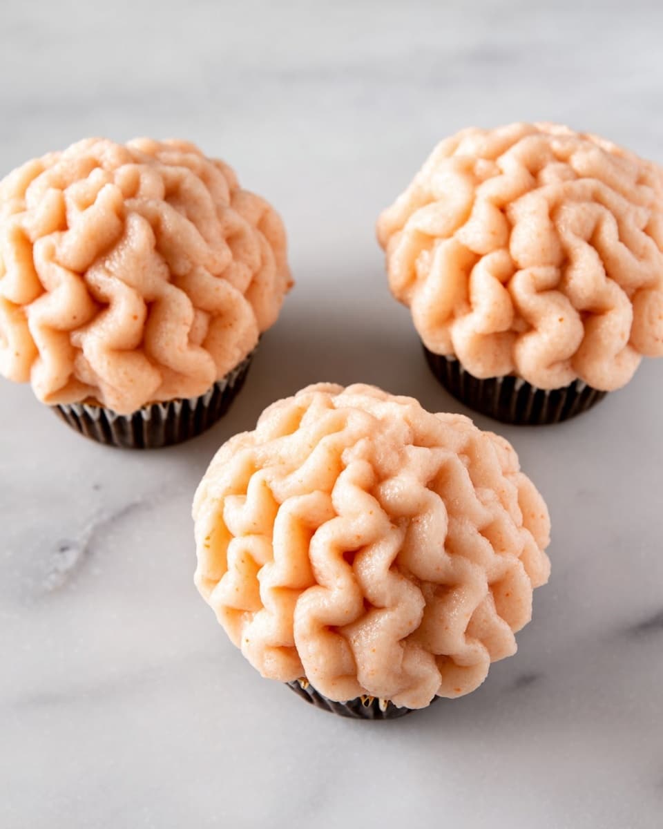 Three cupcakes are shown on a white marbled surface, each topped with a single thick layer of light peach-colored frosting piped in a wavy, swirled pattern that covers the entire top completely. The cupcakes sit in dark brown liners, creating a contrast with the soft color of the frosting. The texture of the frosting looks smooth but dense, and the cupcakes are arranged in a slightly angled row with some space in between. photo taken with an iphone --ar 4:5 --v 7