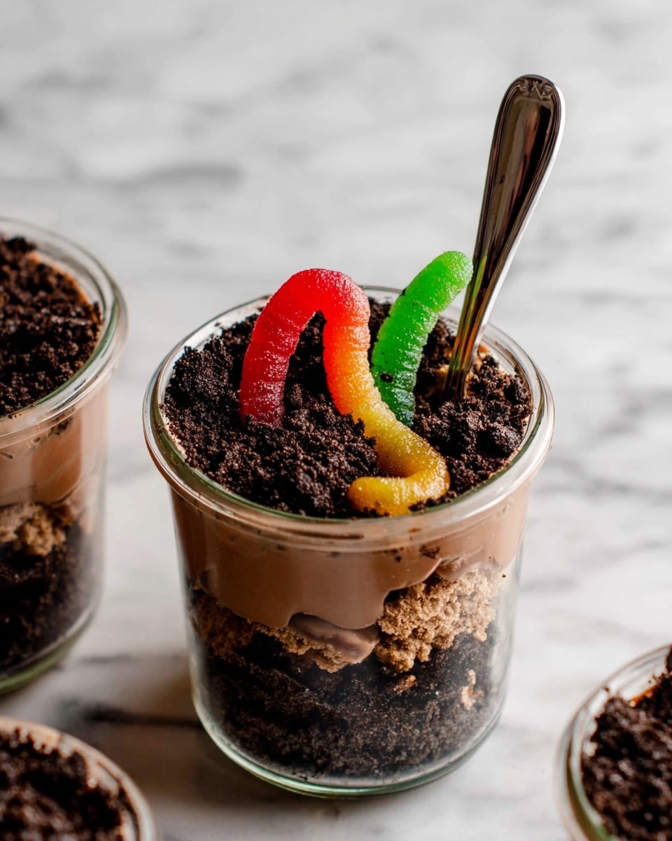 The image shows a clear glass filled with a layered dessert starting with a bottom layer of dark crushed cookie crumbs resembling soil. On top of that is a smooth, creamy chocolate pudding layer, slightly mixed in with some of the cookie crumbs. The dessert is topped with more crushed cookie crumbs to look like dirt, and two colorful gummy worms—one red and green, the other yellow and orange—are placed to appear as if crawling out of the