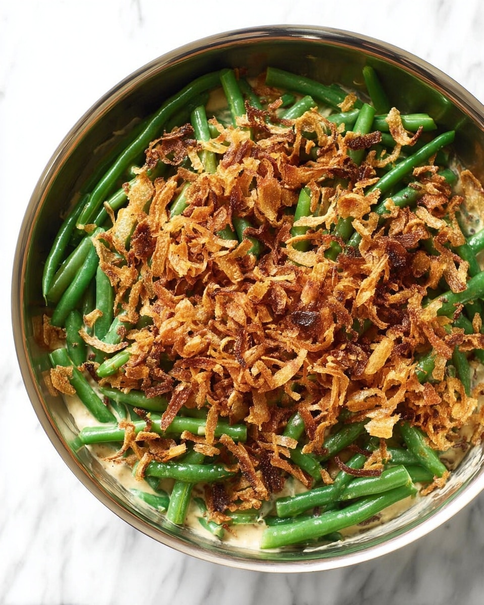 A round metal pan filled with a dish showing three visible layers. The bottom layer is a creamy, light beige sauce partially visible through the green beans layer above. The middle layer consists of fresh green beans, bright green and arranged loosely in the pan. The top layer is a generous, uneven sprinkling of crispy, golden-brown fried onions with a crunchy texture. The background is a white marbled surface. Photo taken with an iphone --ar 4:5 --v 7