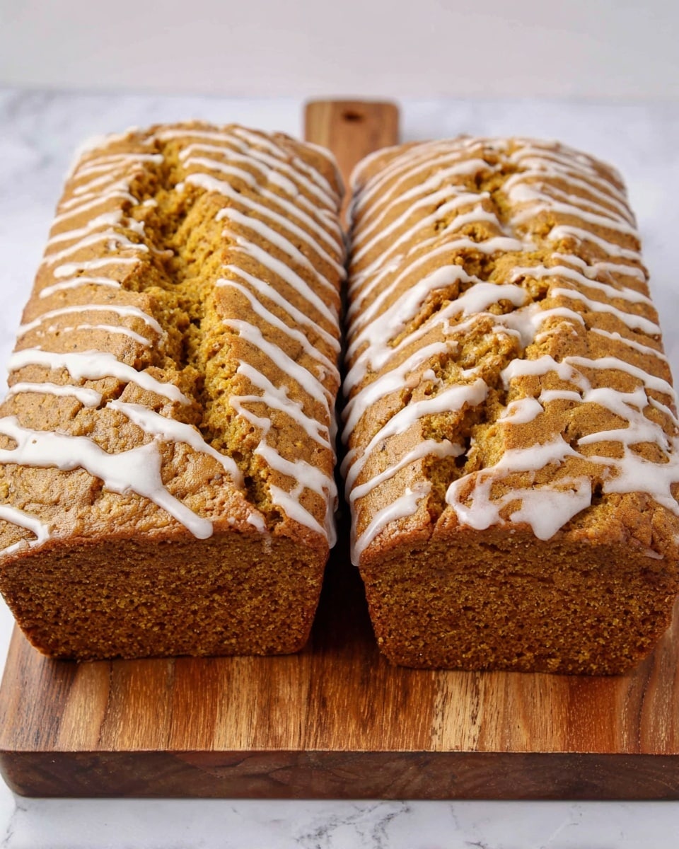 The image shows two loaves of golden brown pumpkin bread placed side by side on a light wooden cutting board over a white marbled surface. The bread has a soft, spongy texture with small holes inside and a lightly cracked crust on top. A thin white icing is drizzled in irregular lines across the top surfaces of both loaves, adding a subtle shine. The loaves are rectangular with rounded edges, and one loaf is shown with a clean cut end facing the viewer, revealing the moist interior. Photo taken with an iphone --ar 4:5 --v 7