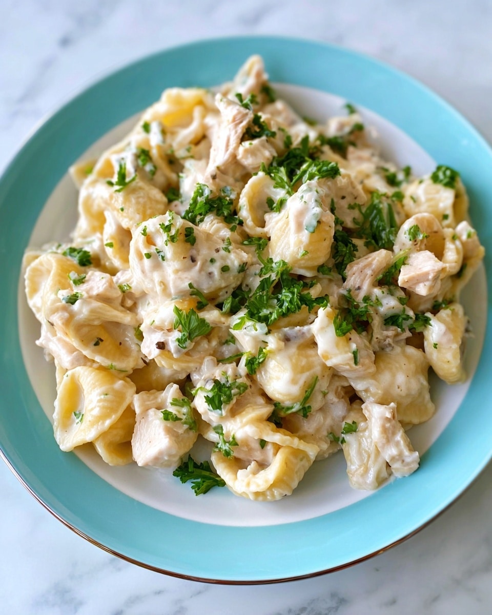 This image shows a white plate with a light blue rim holding a creamy pasta dish. The pasta looks like small folded shells with a pale yellow color, covered with a thick white sauce. Mixed in are small bits of light-colored chicken and finely chopped green parsley sprinkled on top. The sauce glistens slightly, making the dish look rich and smooth, while the parsley adds a fresh green contrast. The plate is placed on a white marbled surface. Photo taken with an iphone --ar 4:5 --v 7
