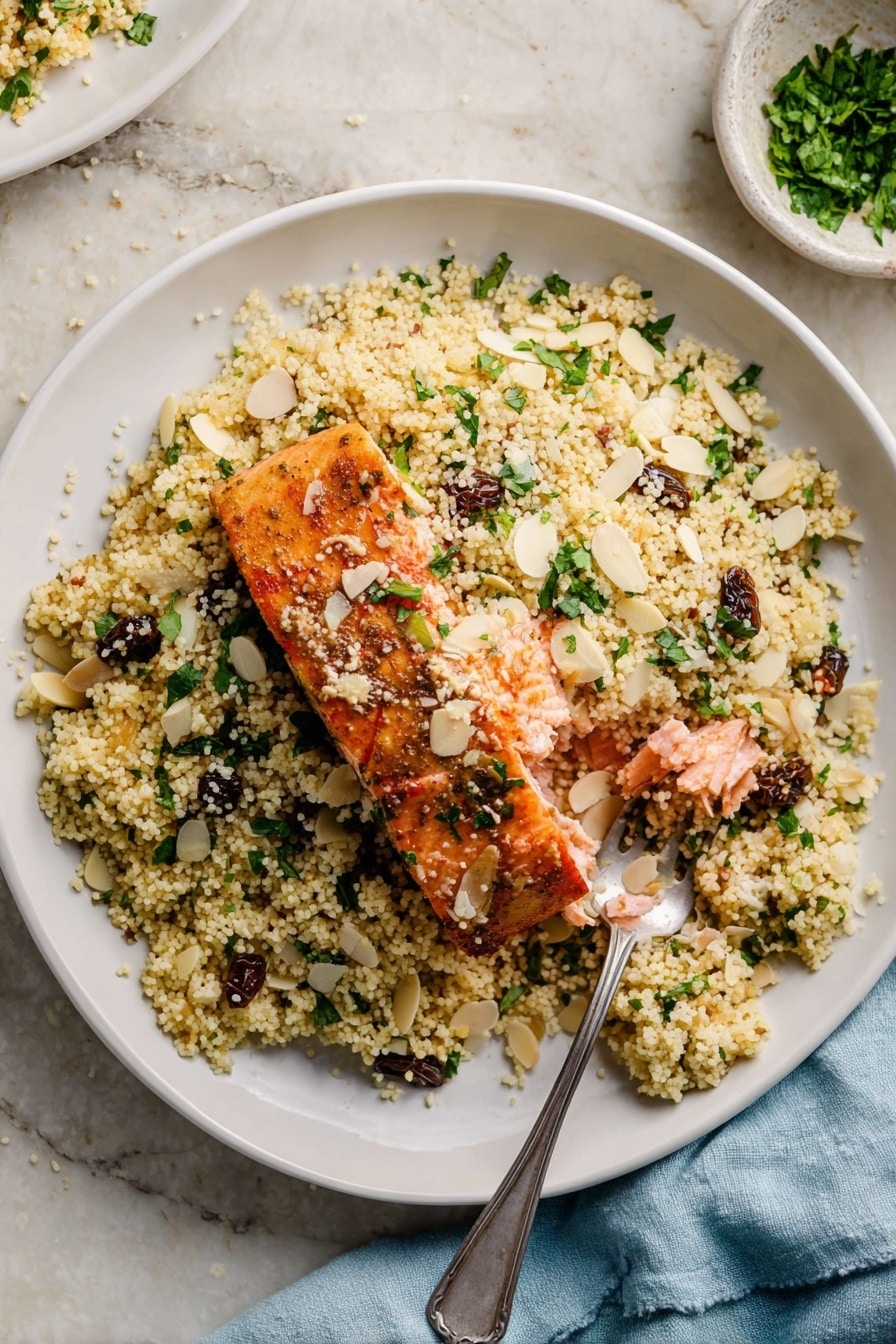 A white bowl filled with a base layer of light yellow couscous mixed with chopped green herbs and small dark raisins. On top of the couscous, there is a piece of cooked salmon with a golden brown crust, broken open to show the soft pink inside. Thin almond slices and extra chopped green herbs are scattered on the dish. A silver spoon rests on the right side of the bowl, partially under the couscous. The bowl sits on a white marbled surface, and a light blue cloth napkin is visible nearby. photo taken with an iphone --ar 2:3 --v 7 - Moroccan-Spiced Salmon and Couscous, quick salmon dinner, Moroccan cuisine recipes, healthy seafood recipes, flavorful fish and couscous