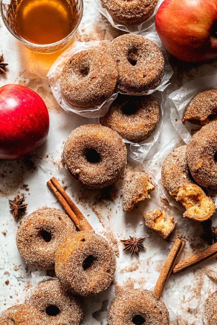 The image shows many cinnamon sugar donuts placed closely on a white marbled surface. Most donuts are whole, with a rough texture covered in sugar and cinnamon. A few donuts are broken, revealing a soft, golden inside with a slightly crumbly texture. Some donuts are wrapped in thin, translucent white paper, adding layers and depth. Near the donuts, there is a bright red apple and a clear glass cup filled with light brown tea and cinnamon sticks inside. Star anise is scattered around for decoration. The scene looks warm and cozy with soft natural light. Photo taken with an iphone --ar 2:3 --v 7 - Baked Cinnamon Crunch Apple Cider Doughnuts, apple cider doughnuts, cinnamon crunch doughnuts, baked fall doughnuts, autumn apple desserts