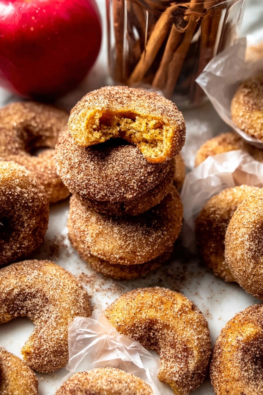 The image shows several crescent-shaped cookies covered in a coarse layer of cinnamon sugar. The cookies are golden brown with a slightly rough texture beneath the sugar coating. Some cookies are stacked in a loose pile, while others are individually wrapped in translucent white paper. The cookies have a crumbly look, with one cookie broken open to reveal a soft yellow interior. In the background, there is a bright red apple and a clear glass jar filled with more cookies and cinnamon sticks on top. The setting uses a white marbled surface underneath all the items. photo taken with an iphone --ar 2:3 --v 7 - Baked Cinnamon Crunch Apple Cider Doughnuts, apple cider doughnuts, cinnamon crunch doughnuts, baked fall doughnuts, autumn apple desserts