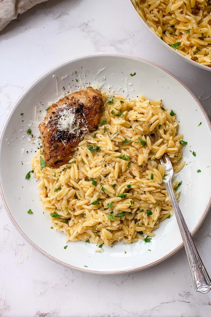 A white plate with a serving of golden pasta mixed with small green parsley pieces and bits of light brown toasted orzo, covering most of the plate's center and right side. On the left side of the plate, there is a browned, cooked piece of chicken skin-on, topped lightly with grated white cheese and some black pepper sprinkled over both pasta and chicken. A silver fork rests diagonally on the plate, partly under the pasta on the right side. The plate sits on a white marbled surface, and part of a bowl of similar pasta is visible in the top right corner. Photo taken with an iphone --ar 2:3 --v 7 - One Pot Garlic Butter Chicken and Orzo, easy one pot chicken dinner, garlic butter chicken thighs, orzo pasta recipes, quick weeknight chicken meal