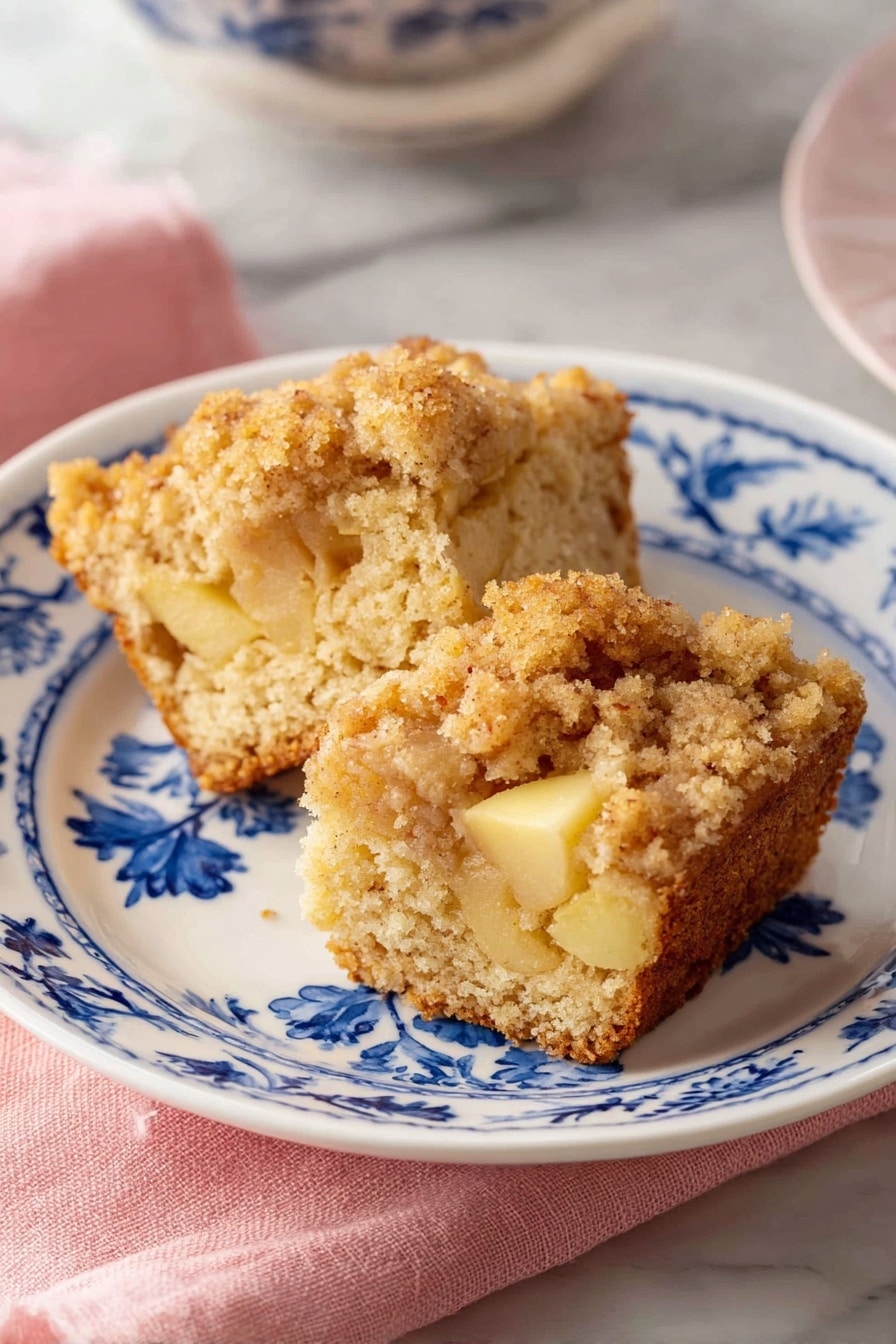 Two pieces of crumb-topped cake are placed on a white plate with blue floral patterns. The cake has a light brown color with soft, crumbly texture and visible chunks of yellow apple inside. The crumb topping is thicker and golden brown. The plate sits on a pink cloth, all set on a white marbled surface. Photo taken with an iphone --ar 2:3 --v 7 - Apple Muffins, apple muffins recipe, moist apple muffins, cinnamon apple muffins, easy breakfast muffins