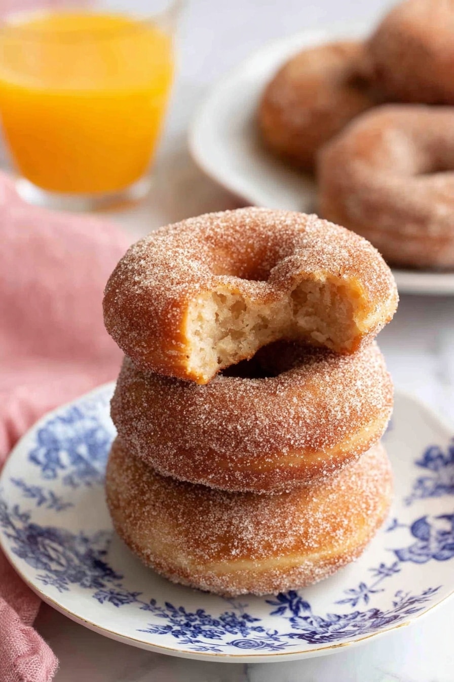 Three cinnamon sugar donuts are stacked on a white plate with blue floral designs, showing a soft texture covered in fine sugar crystals; the top donut has a bite taken, revealing a light, fluffy inside with a slightly darker core. Behind the plate is a blurred white plate with more cinnamon sugar donuts and a clear glass filled with bright orange juice, all set on a white marbled surface. A pink cloth is placed softly on the left side. photo taken with an iphone --ar 2:3 --v 7 - Apple Cider Donuts, autumn donut recipes, cozy fall treats, crispy baked donuts, homemade apple cider donuts