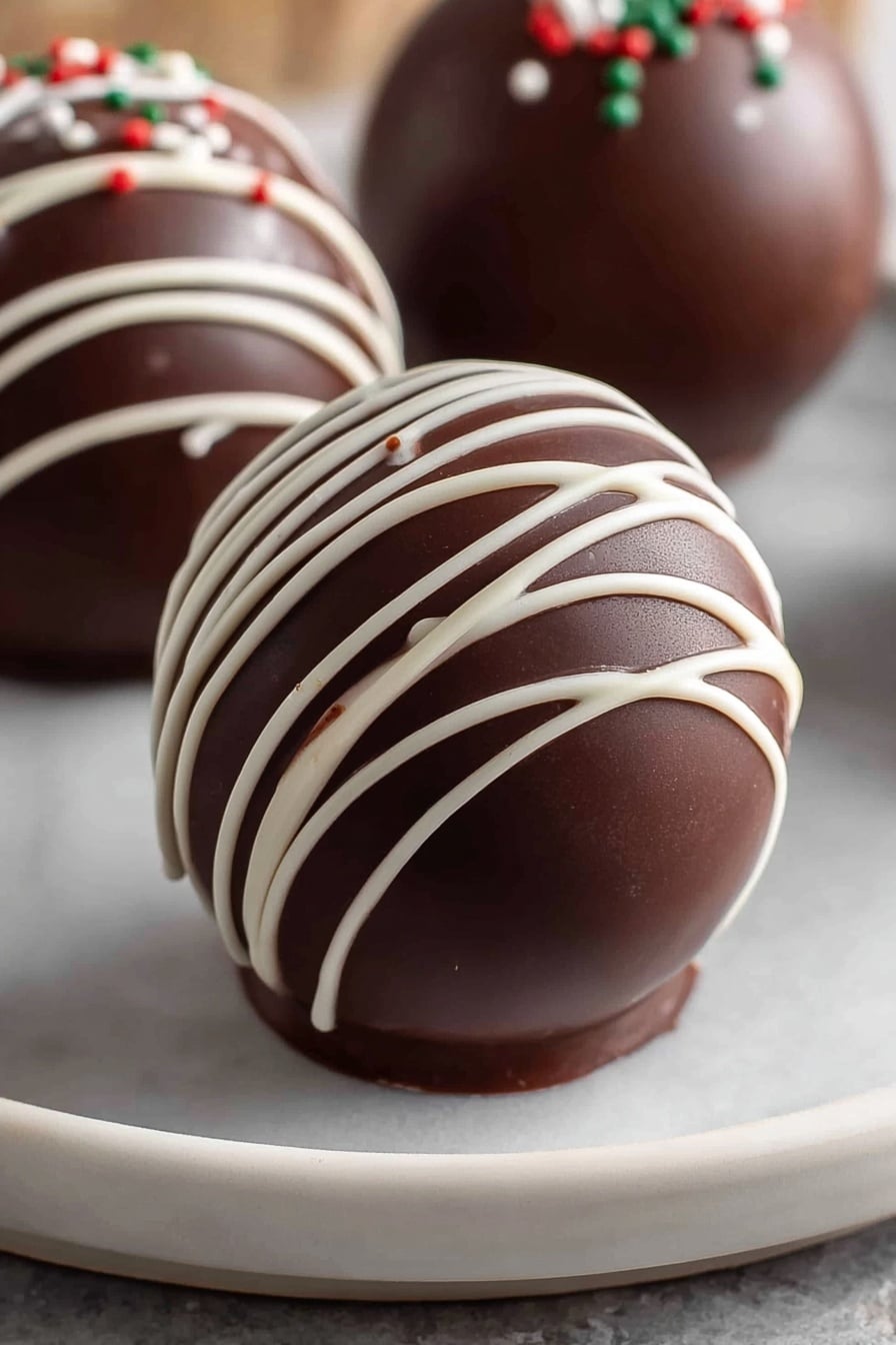 The image shows a smooth, shiny, dark brown chocolate sphere placed on a white plate with a white marbled texture under it. The sphere has thin white chocolate lines drizzled across its surface in a loose crisscross pattern, creating contrast and delicate texture on the curved chocolate shell. In the background, two more similar chocolate spheres are partially visible, each decorated with the same white drizzle and small dots of colored sprinkles. The overall look is clean and glossy, with the sphere’s curved layers clearly defined. photo taken with an iphone --ar 2:3 --v 7 - Delicious Hot Chocolate Bombs, Hot Chocolate Bombs recipe, homemade hot chocolate bombs, easy hot chocolate bombs, festive hot chocolate drinks