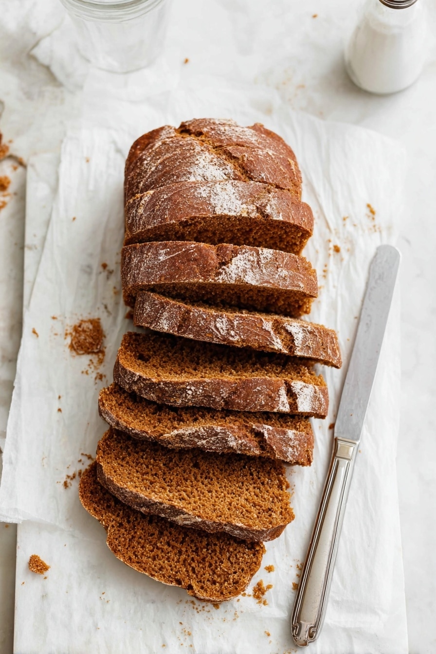 The image shows a loaf of brown bread cut into six thick slices, placed slightly overlapping each other on a white parchment paper. The bread has a textured crust with a few light flour spots on top and a soft, crumbly inside with a warm brown color. To the right of the bread, there is a silver knife with a simple handle resting on the parchment paper. The background is a white marbled surface with some bread crumbs scattered around, and the top right part of a clear glass and a white salt shaker are visible. Photo taken with an iphone --ar 2:3 --v 7 - Easy Moist Pumpkin Bread, fall pumpkin bread, moist pumpkin bread recipe, pumpkin bread with spices, soft pumpkin quick bread