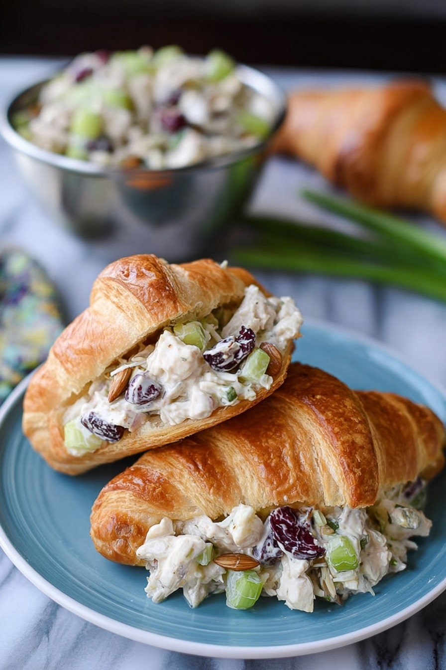 The image shows two croissants on a blue plate placed on a white marbled surface. One croissant is whole with a golden-brown, flaky texture. The other croissant is open and filled with a creamy salad made of small white chicken pieces, chopped celery, dried dark cranberries, sunflower seeds, and light beige dressing, layered inside the croissant’s light and flaky interior. In the background, there is a metal bowl filled with more of the salad, and some green onions and an additional croissant are blurred. Photo taken with an iphone --ar 2:3 --v 7 - Cranberry Chicken Salad, cranberry chicken salad recipe, healthy chicken salad with cranberries, easy chicken salad with cranberries, fruit chicken salad recipe