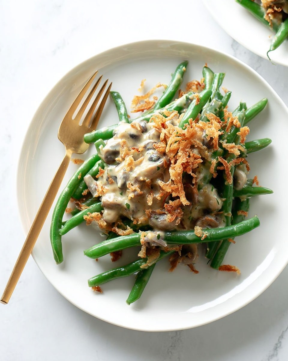 A white plate holds a serving of fresh green beans topped with a creamy beige sauce mixed with small bits of mushrooms. The beans are arranged in a loose pile, with crispy golden fried onions sprinkled on top, adding texture. A gold fork rests on the left side of the plate, its handle extending outward. The surface beneath the plate is a white marbled texture. photo taken with an iphone --ar 4:5 --v 7