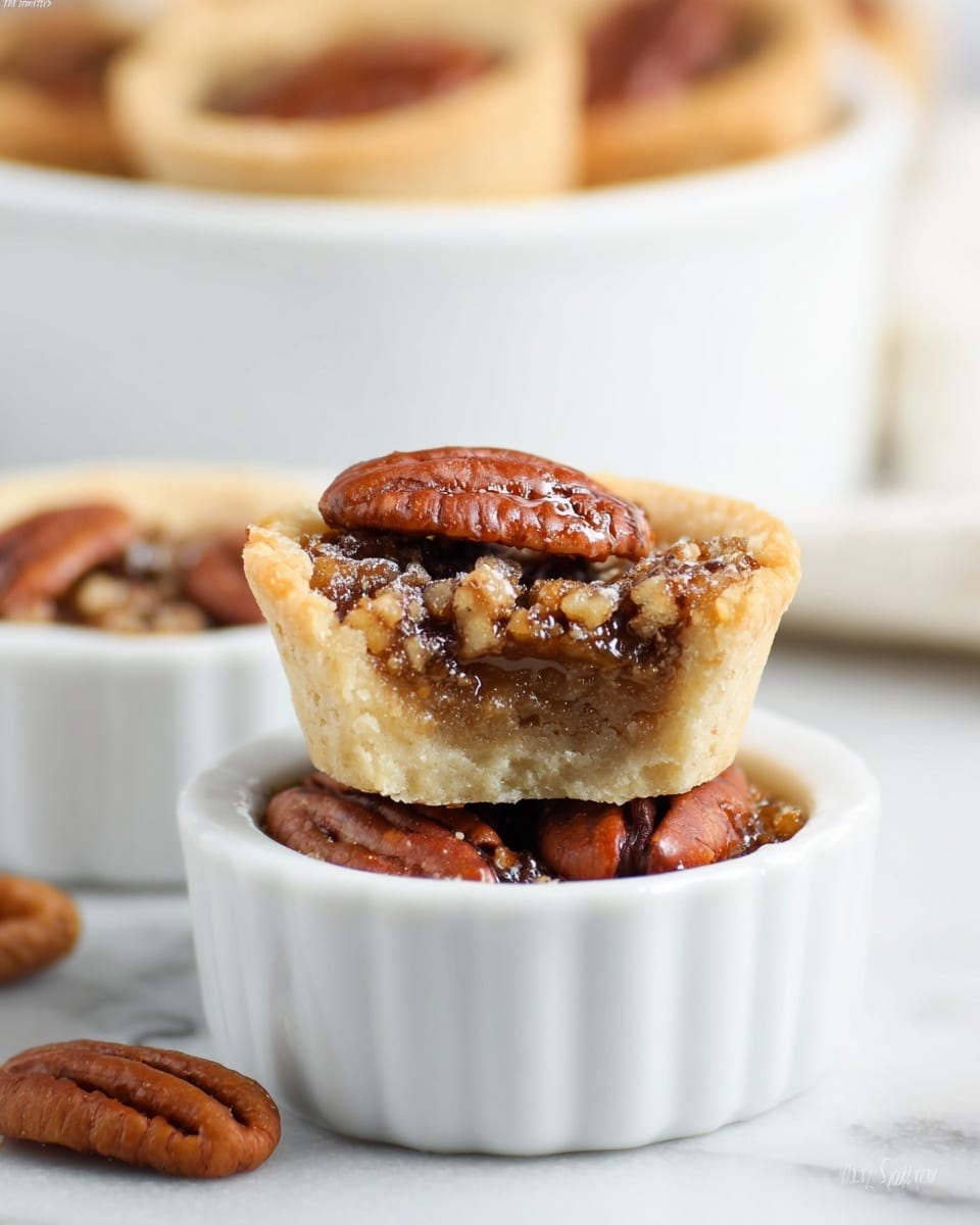 A small white ramekin filled with several smooth, brown pecan halves sits on a white marbled surface. On top, resting slightly tilted, is a mini tart with a pale, crumbly crust forming the base and sides. Inside the tart is a textured, moist filling in a deep golden brown color with shiny spots, suggesting melted chocolate or syrup mixed with chopped pecans. In the blurred background, a white serving dish holds more of these mini tarts stacked together. Photo taken with an iphone --ar 4:5 --v 7