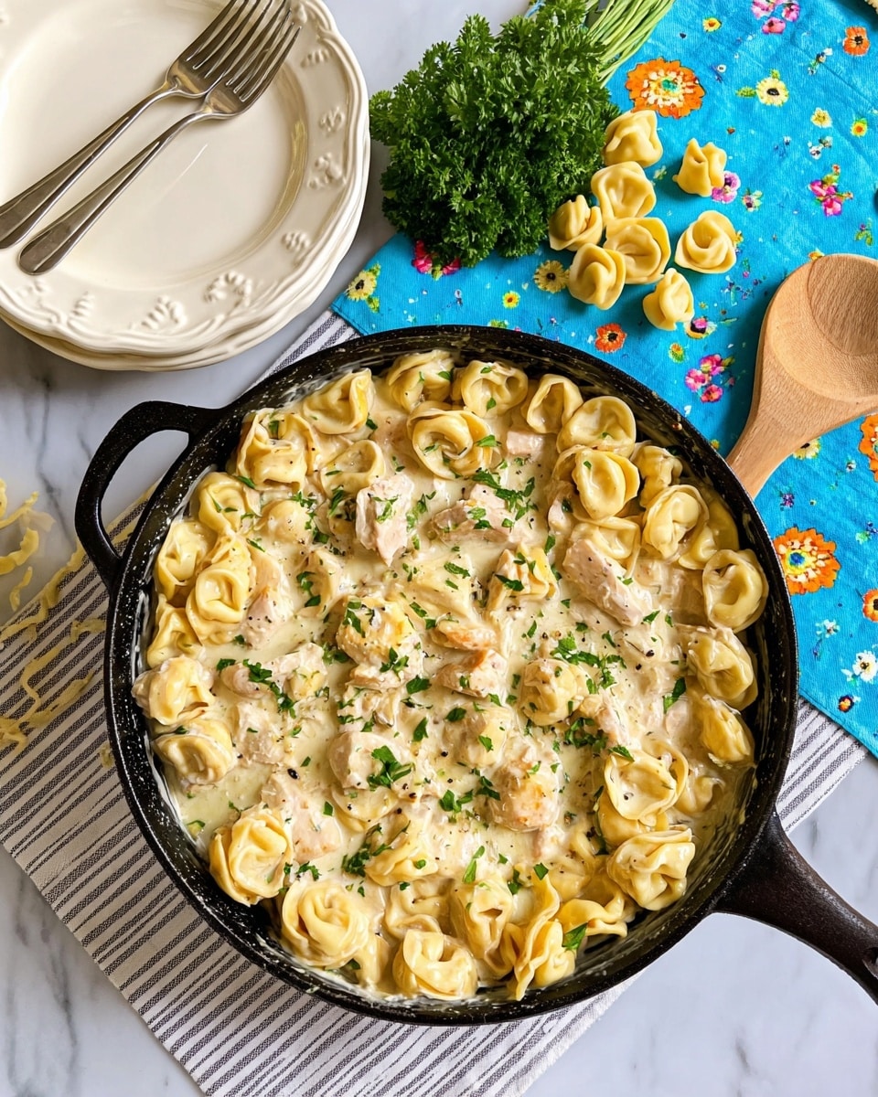 A black cast iron pan filled with creamy tortellini pasta in a white sauce mixed with pieces of light-colored chicken and sprinkled with small green parsley leaves on top, sitting on a white marbled surface covered by a striped cloth. To the left of the pan, there are two stacked white plates with a scalloped edge, and two forks crossed on the top plate. A bunch of green parsley and uncooked tortellini pieces are placed near the top edge, while to the right is a wooden spoon resting on a blue cloth with small colorful flowers. Photo taken with an iphone --ar 4:5 --v 7
