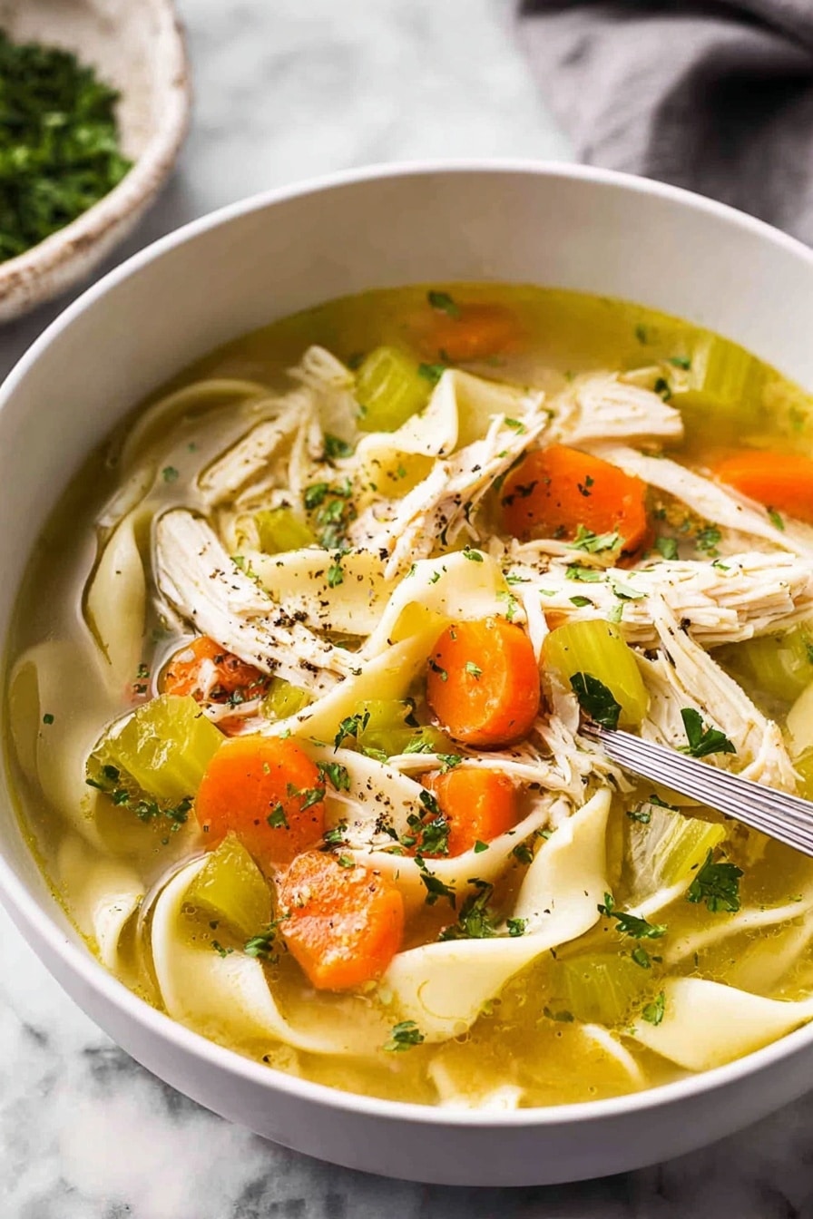 The image shows a close-up view of a white bowl filled with clear chicken soup. Inside the soup, there are wide flat noodles curled softly, bright orange carrot slices, pieces of pale green celery, and shredded white chicken meat scattered on one side. Small green parsley flakes float on the surface along with some black pepper specks. The soup is light golden and clear, with visible layers of ingredients resting naturally in the broth. The bowl sits on a white marbled surface. photo taken with an iphone --ar 2:3 --v 7 - Homemade Chicken Noodle Soup from Scratch, homemade chicken noodle soup, scratch-made chicken soup, homemade chicken soup recipe, comforting chicken noodle soup