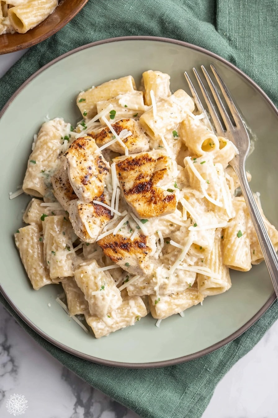 A white plate holds a serving of rigatoni pasta coated in a creamy white sauce with small green herb flecks. On top of the pasta lie four grilled chicken pieces, light brown with darker grill marks, partially covered by the sauce. Thin white shredded cheese is sprinkled over the chicken and pasta, adding texture. A silver fork rests on the right side of the plate. The plate is on a soft green cloth placed on a white marbled surface. Photo taken with an iphone --ar 2:3 --v 7 - One Pot Creamy Parmesan Chicken Pasta, creamy chicken pasta, one pot pasta dinner, comforting chicken pasta recipe, easy cheesy chicken pasta