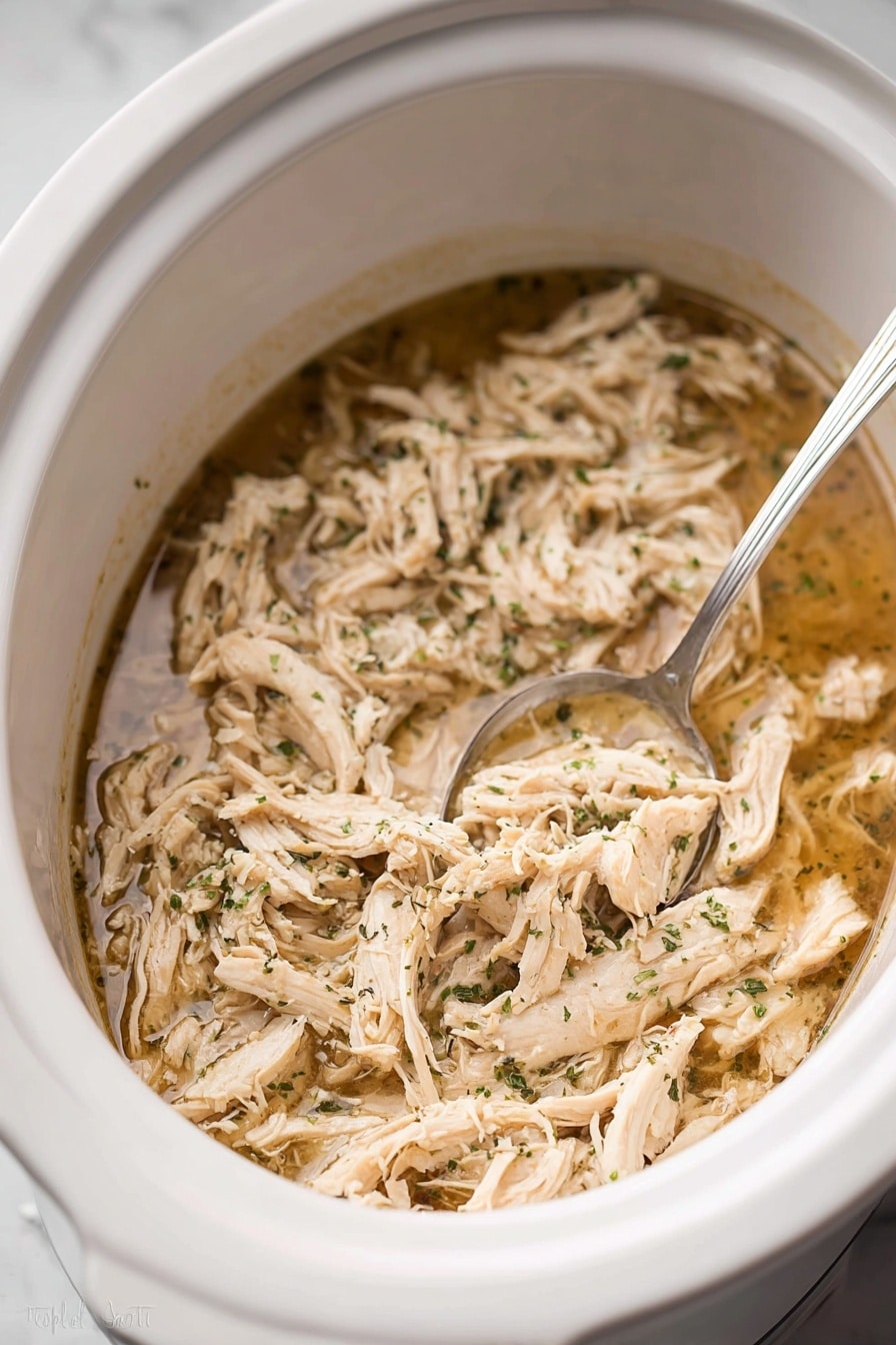 Inside a white slow cooker, there is one main layer of shredded light beige cooked chicken mixed with small green herb pieces, all soaked in a thin, light brown broth. A silver spoon rests partially submerged in the mixture on the right side. The chicken strands have a soft texture, and the herbs are finely sprinkled. The slow cooker sits on a white marbled surface. photo taken with an iphone --ar 2:3 --v 7 - Juicy Crockpot Shredded Chicken, easy shredded chicken, slow cooker chicken recipe, tender chicken for meal prep, versatile shredded chicken