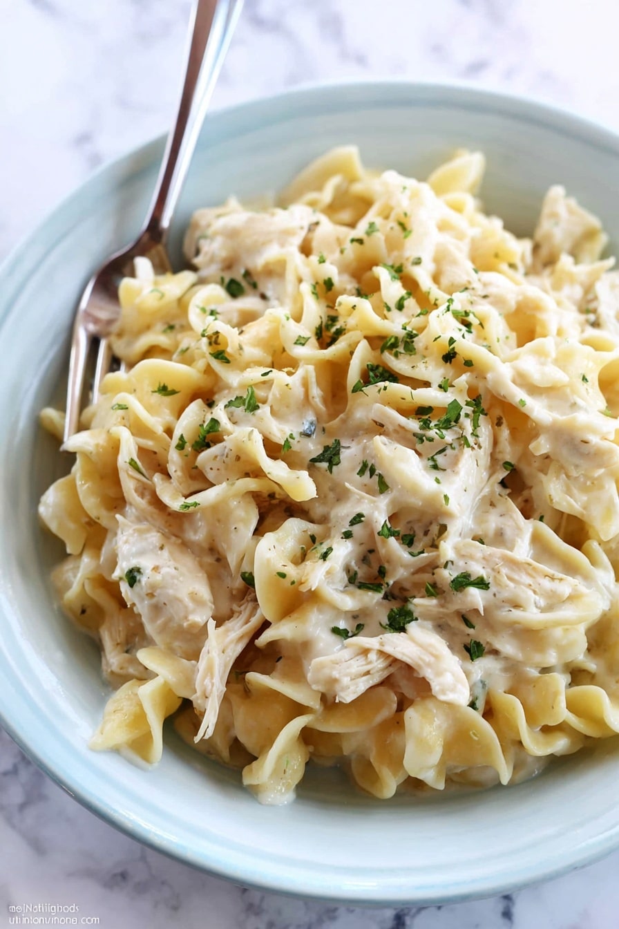 A close-up of creamy chicken noodle dish served in a white bowl placed on a white marbled surface. The dish has three main layers: at the bottom is a thick white cream sauce, on top of it a layer of soft cooked egg noodles with a slightly wavy texture and pale yellow color, and scattered throughout and on top are pieces of shredded chicken mixed well with the noodles. The whole dish is sprinkled with small green herb flakes for garnish. A fork is resting inside the bowl, ready to eat. photo taken with an iphone --ar 2:3 --v 7 - Crockpot Chicken and Noodles, slow cooker chicken and noodles, easy chicken noodle dinner, comforting slow cooker recipes, family-friendly crockpot meals