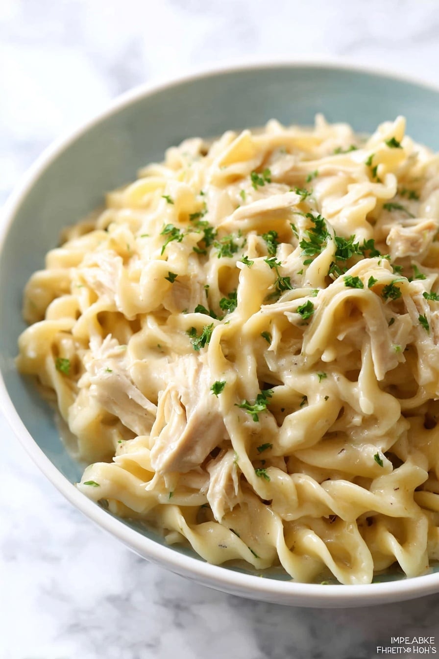 A close-up of a white bowl filled with creamy egg noodles mixed with shredded pieces of light beige chicken. The egg noodles are thick and twisted, coated in a smooth, light cream sauce. Small bits of green herbs are sprinkled on top and some are mixed within the noodles, adding small dots of green color. The bowl is set on a white marbled surface, creating a clean and bright background. Photo taken with an iphone --ar 2:3 --v 7 - Crockpot Chicken and Noodles, slow cooker chicken and noodles, easy chicken noodle dinner, comforting slow cooker recipes, family-friendly crockpot meals