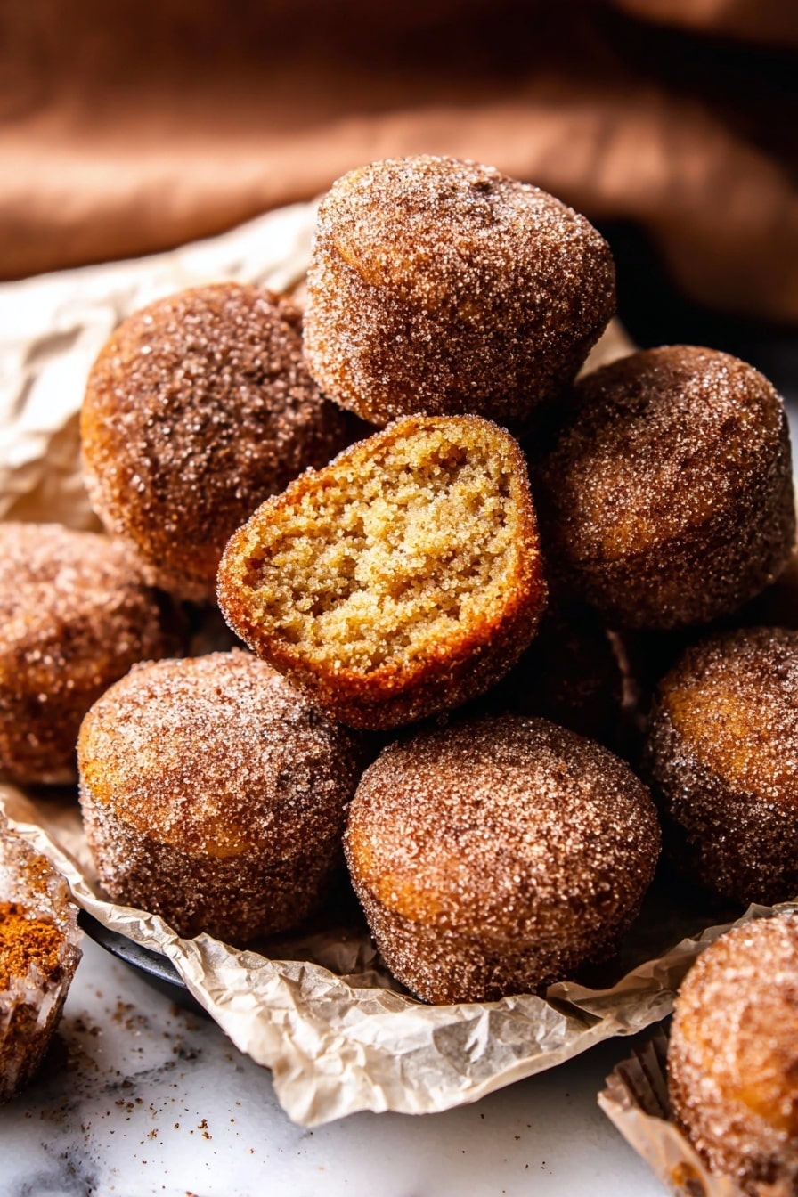 The image shows a pile of small, round doughnut muffins covered in cinnamon sugar on a white marbled surface. There are about ten muffins closely stacked, with one muffin on top showing its inside layer which is soft and light brown with a slightly crumbly texture. The muffins have a rough, grainy outer layer from the sugar coating, and they are resting on crumpled white parchment paper. The scene captures a warm, cozy feeling from the brown and golden hues of the muffins, with some muffins sitting inside a black tray. photo taken with an iphone --ar 2:3 --v 7 - Baked Cinnamon Sugar Apple Cider Doughnut Holes, apple cider doughnut holes, baked fall donut bites, cinnamon sugar donut holes, cozy fall treats