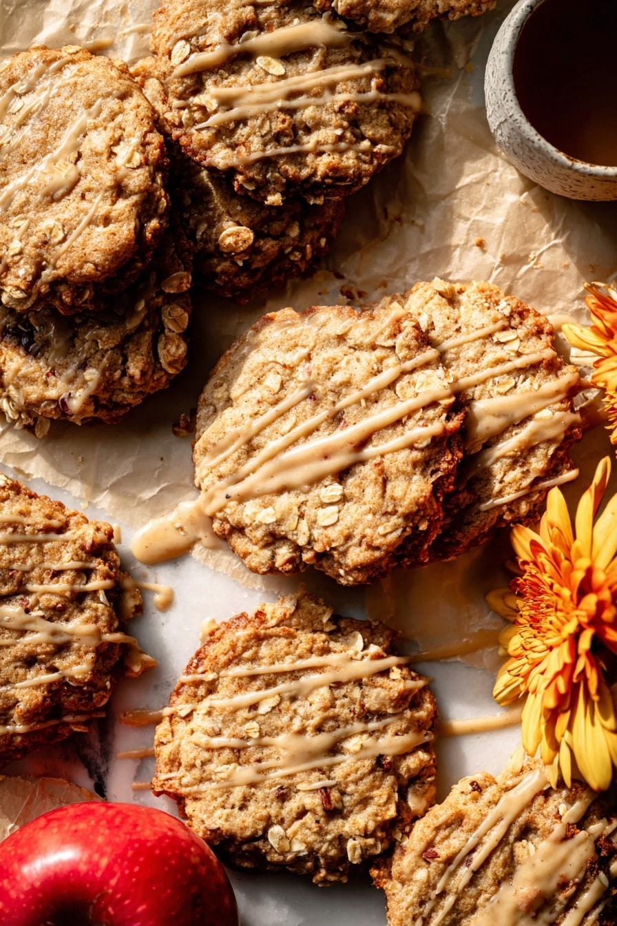 Several oatmeal cookies are spread on light brown parchment paper over a white marbled surface. The cookies are golden brown with a rough texture, showing visible oats and slightly darker edges that look crispy. Each cookie is drizzled with a thin layer of light caramel-colored glaze, creating irregular lines and patches across their tops. In the bottom left corner of the image, two whole red apples with light yellow patches are placed together, adding a fresh contrast to the warm cookie colors. The light source creates soft shadows and highlights, enhancing the cookies’ textures and the glaze's shine. photo taken with an iphone --ar 2:3 --v 7 - Iced Apple Cider Caramel Oatmeal Cookies, fall-flavored oatmeal cookies, caramel apple cookies, cozy autumn cookie recipe, easy fall cookie ideas