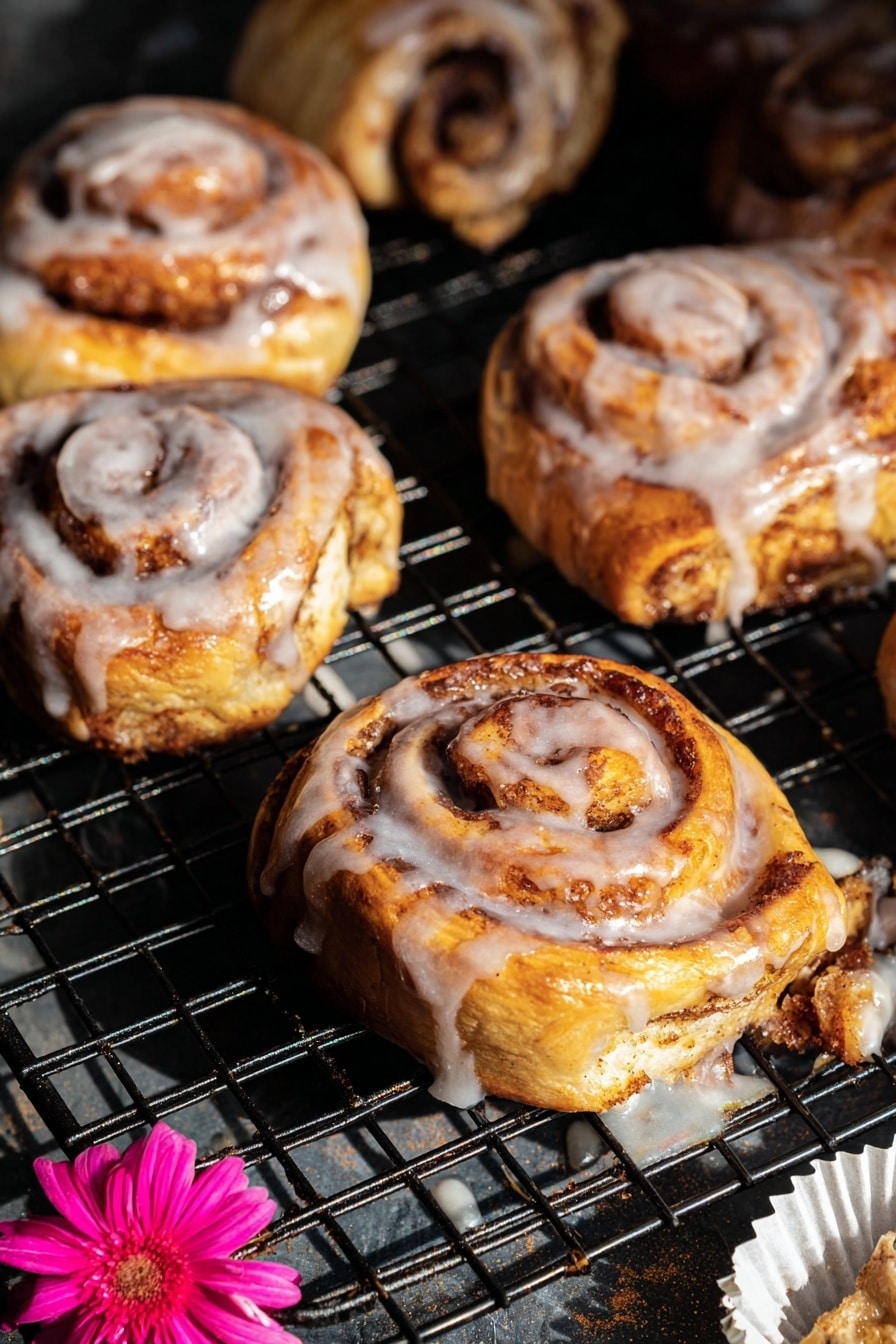 The image shows several cinnamon rolls on a black wire cooling rack with a dark base underneath. Each cinnamon roll has visible spiral layers of golden-brown dough swirled with darker brown cinnamon sugar. The top layers are coated with a shiny white glaze that drips slightly down the sides and onto the rack. The rolls have a soft, fluffy texture with a mix of light yellow and brown tones, and some darker, slightly crisped edges. Near the bottom right, there is a white paper liner with a bright pink flower placed on it. The light creates highlights on the glossy glaze and shadows between the cinnamon rolls, giving the scene depth and warmth. Photo taken with an iphone --ar 2:3 --v 7 - Shortcut Easy Apple Fritters, apple fritters using puff pastry, quick apple fritters, cinnamon apple bites, fall dessert recipes