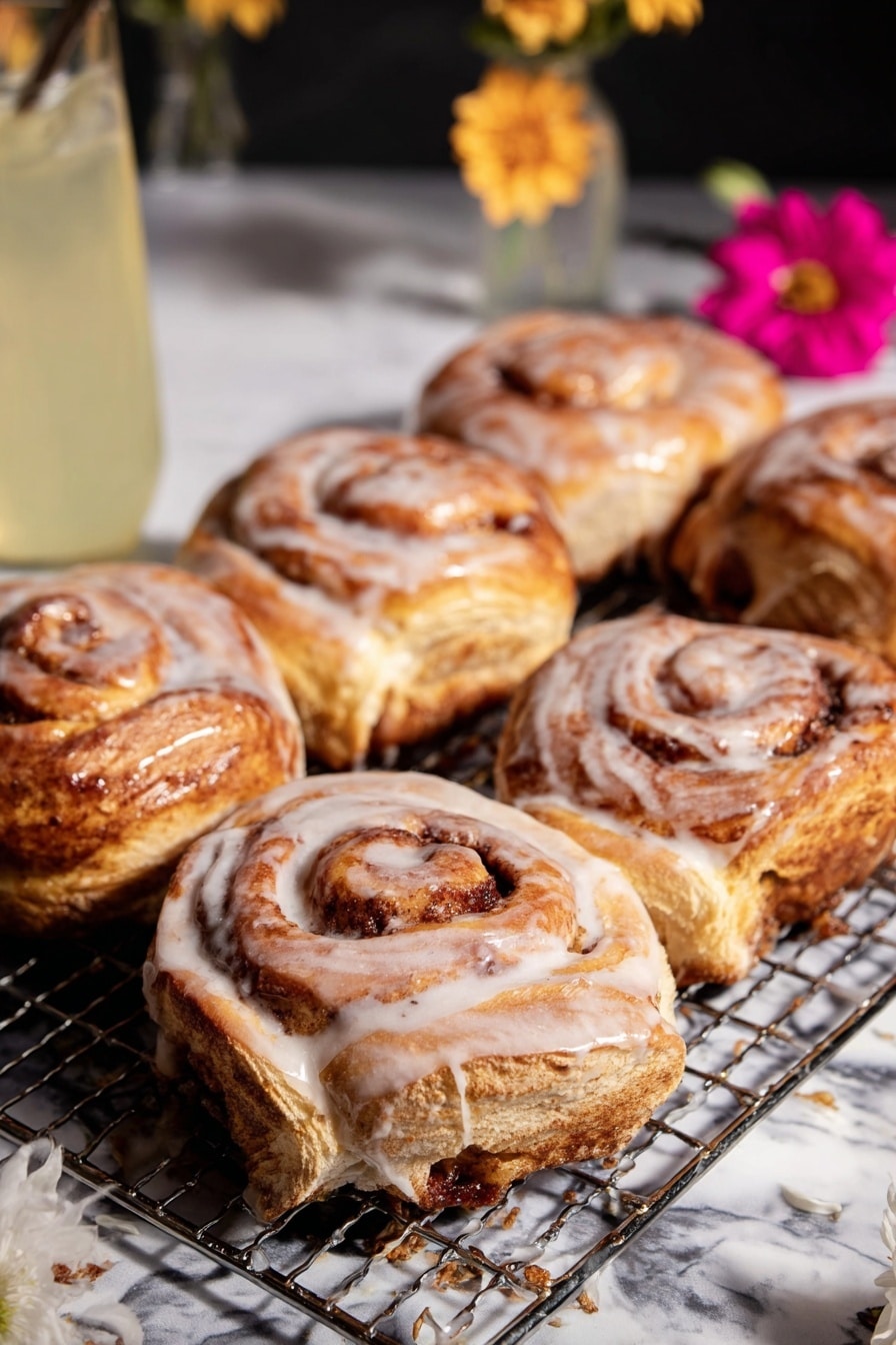 The image shows six cinnamon rolls with a thick white glaze, placed on a wire cooling rack over a white marbled surface. Each cinnamon roll has multiple golden brown layers swirled tightly together with visible cinnamon filling, some layers darker due to caramelized sugar. The glaze drips slightly down the sides, adding a shiny texture on top of the soft, fluffy layers. A white paper liner holds one cinnamon roll in the front, surrounded by a few splashes of glaze on the surface. In the background, a tall glass with a light yellow liquid and a flower in a small white vase add a soft decorative touch, along with a pink flower in a white paper liner near the lower right corner. photo taken with an iphone --ar 2:3 --v 7 - Shortcut Easy Apple Fritters, apple fritters using puff pastry, quick apple fritters, cinnamon apple bites, fall dessert recipes