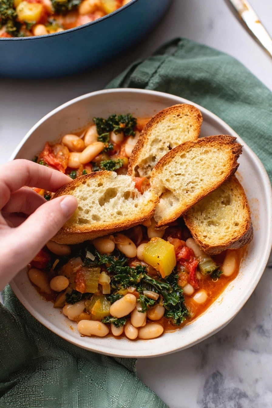 A white bowl filled with a warm stew made of white beans, chunks of yellow squash, wilted green kale, and small red tomato pieces. On top, three slices of toasted baguette bread rest, one slice held by a woman's hand, showing the mix of beans and vegetables piled on it. The stew's sauce is a light red color, and the bread has a golden-brown crust with a porous, soft inside texture. The bowl sits on a green cloth on a white marbled surface, with part of a blue pot filled with more stew blurred in the background. Photo taken with an iphone --ar 2:3 --v 7 - Stewed Great Northern Beans with Kale, Tomatoes, and Herbs, healthy bean and kale stew, hearty vegetarian dinner, easy bean recipes, plant-based comfort food