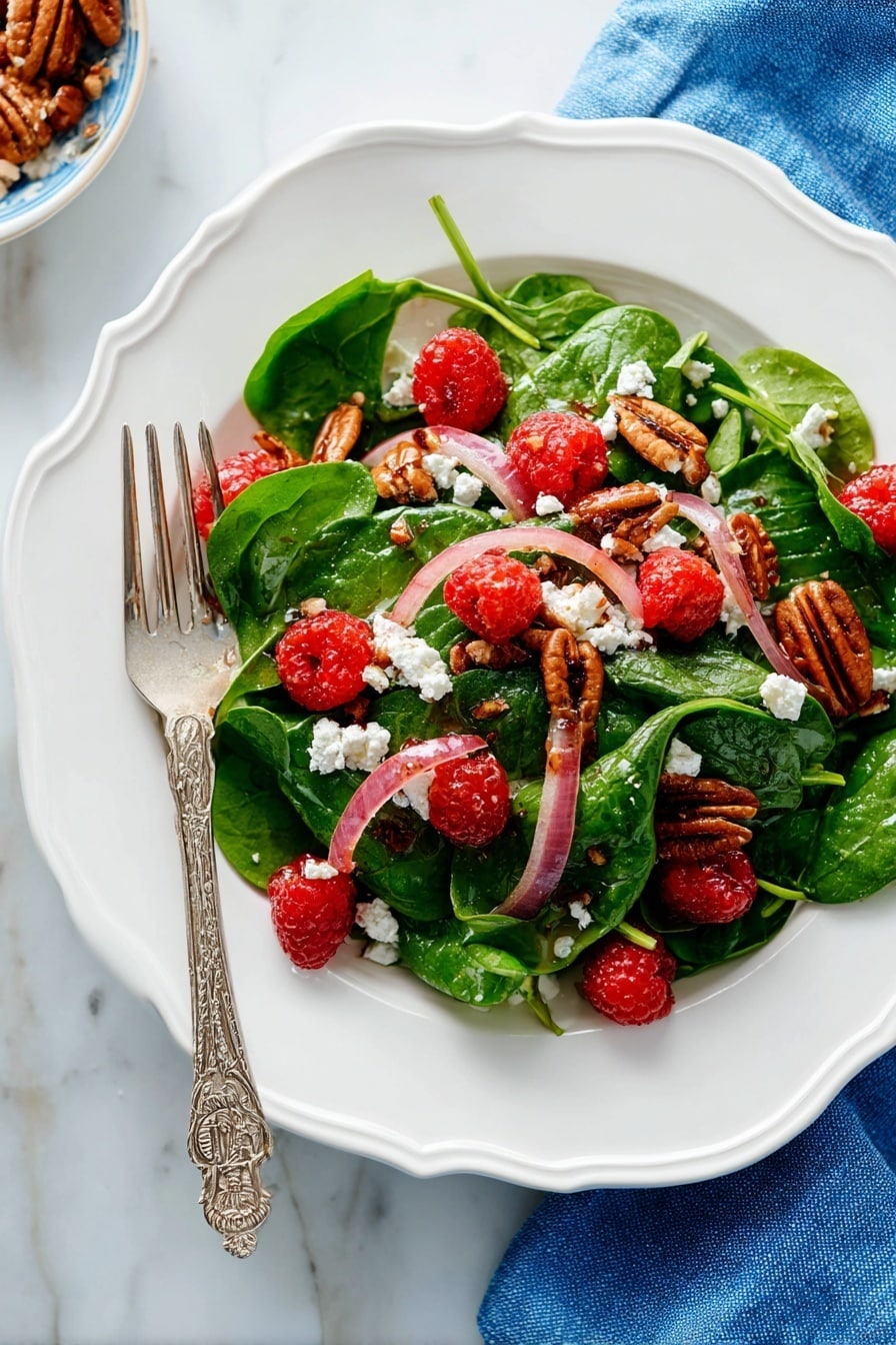 A white scalloped plate holds a fresh salad with three main layers: the base layer has bright green spinach leaves with soft texture, the middle layer has thin slices of pale pink-red onions scattered evenly, and the top layer features bright red raspberries, crumbled white cheese, and brown pecan halves arranged to create color contrast. A silver fork with an ornate handle rests on the left side of the plate, partially on the salad. The plate is placed on a white marbled surface with a blue cloth napkin nearby. Photo taken with an iphone --ar 2:3 --v 7 - Spinach Salad with Goat Cheese Raspberries and Toasted Pecans, healthy spinach salad with goat cheese and berries, quick gourmet spinach salad recipe, fresh berry and pecan salad, easy spinach salad with cheese and nuts