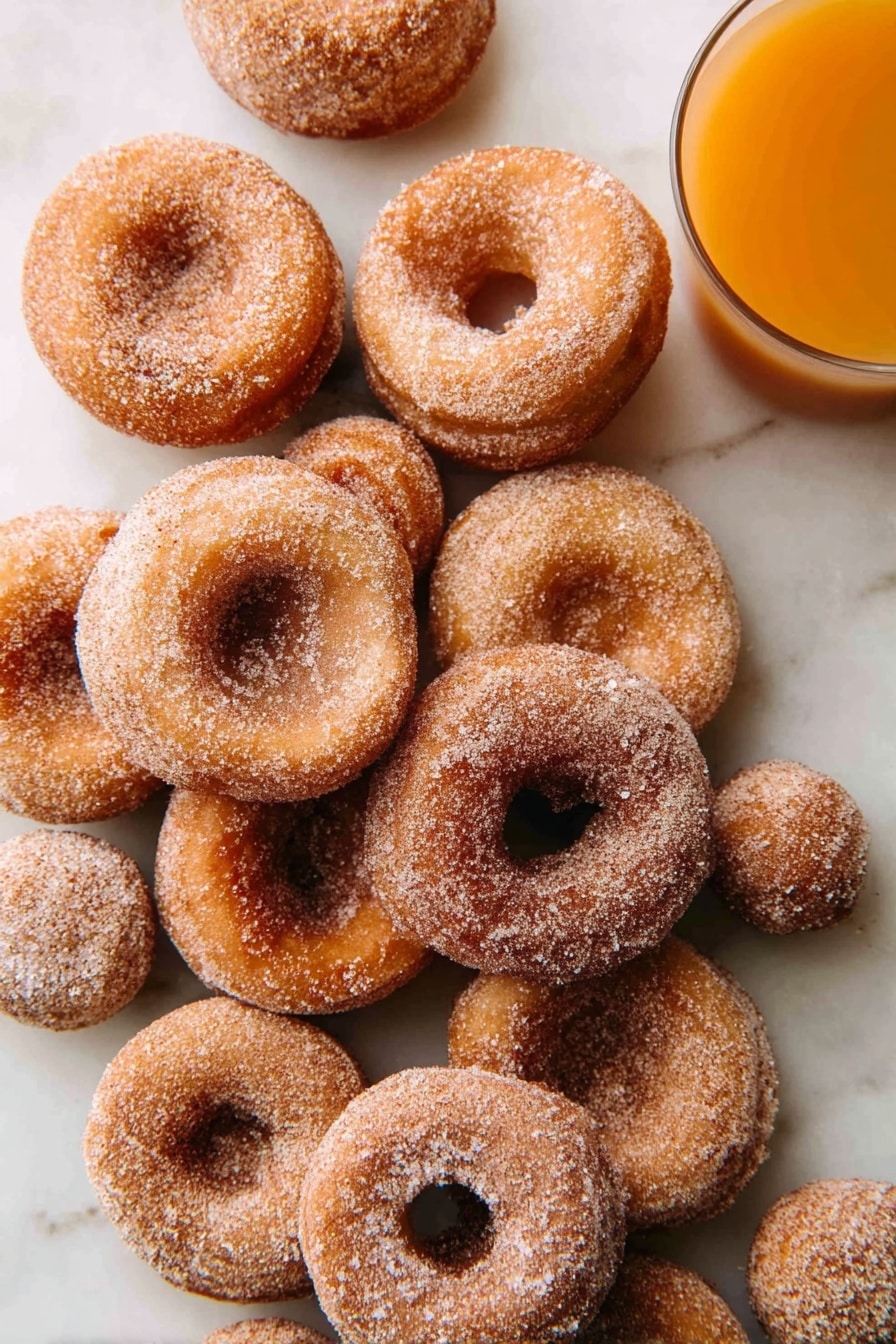 A pile of about a dozen small round donuts with light brown color, coated evenly with granulated sugar and cinnamon, arranged casually on a white marbled surface; two small round donut holes are also visible among the donuts, and at the top right corner, a clear glass of orange-colored juice is partially shown. photo taken with an iphone --ar 2:3 --v 7 - Apple Cider Donuts, autumn donut recipes, cozy fall treats, crispy baked donuts, homemade apple cider donuts