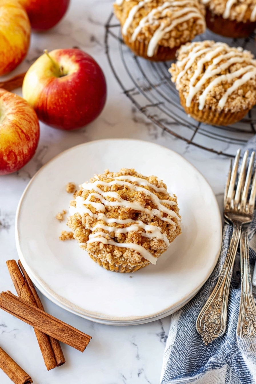 A single round muffin sits in the middle of a white plate, topped with a crumbly golden-brown streusel layer and drizzled with thick white icing in uneven stripes. The plate is on a white marbled surface surrounded by three shiny red and yellow apples, two cinnamon sticks, and a blue and white striped cloth. To the right, a wire cooling rack holds several more muffins with the same crumb topping and white icing drizzle. Two ornate silver forks rest beside the plate. The scene is bright and cozy, with warm autumn colors and textures. photo taken with an iphone --ar 2:3 --v 7 - Cinnamon Apple Muffins with Crumble Topping and Glaze, apple cinnamon muffins, homemade apple muffins, fall breakfast muffins, easy apple muffin recipe
