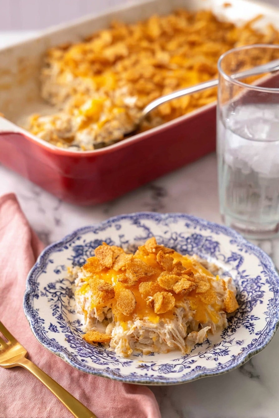 A portion of a creamy casserole is served on a white plate with blue floral patterns, showing three main layers: the bottom layer is shredded white meat mixed with a light creamy sauce, the middle layer has melted orange cheese blended with the meat, and the top layer consists of crushed crispy crackers with more melted cheese giving a golden-brown, crunchy texture. Behind the plate, there is a large red baking dish filled with the same casserole, with a silver serving spoon inside. To the right of the plate is a clear glass of ice water, and to the left is a gold fork resting on a pink cloth napkin, all placed on a white marbled surface. photo taken with an iphone --ar 2:3 --v 7 - Creamy Chicken Casserole, chicken casserole recipes, easy comfort food, cheesy casserole dinner, family-friendly chicken bake