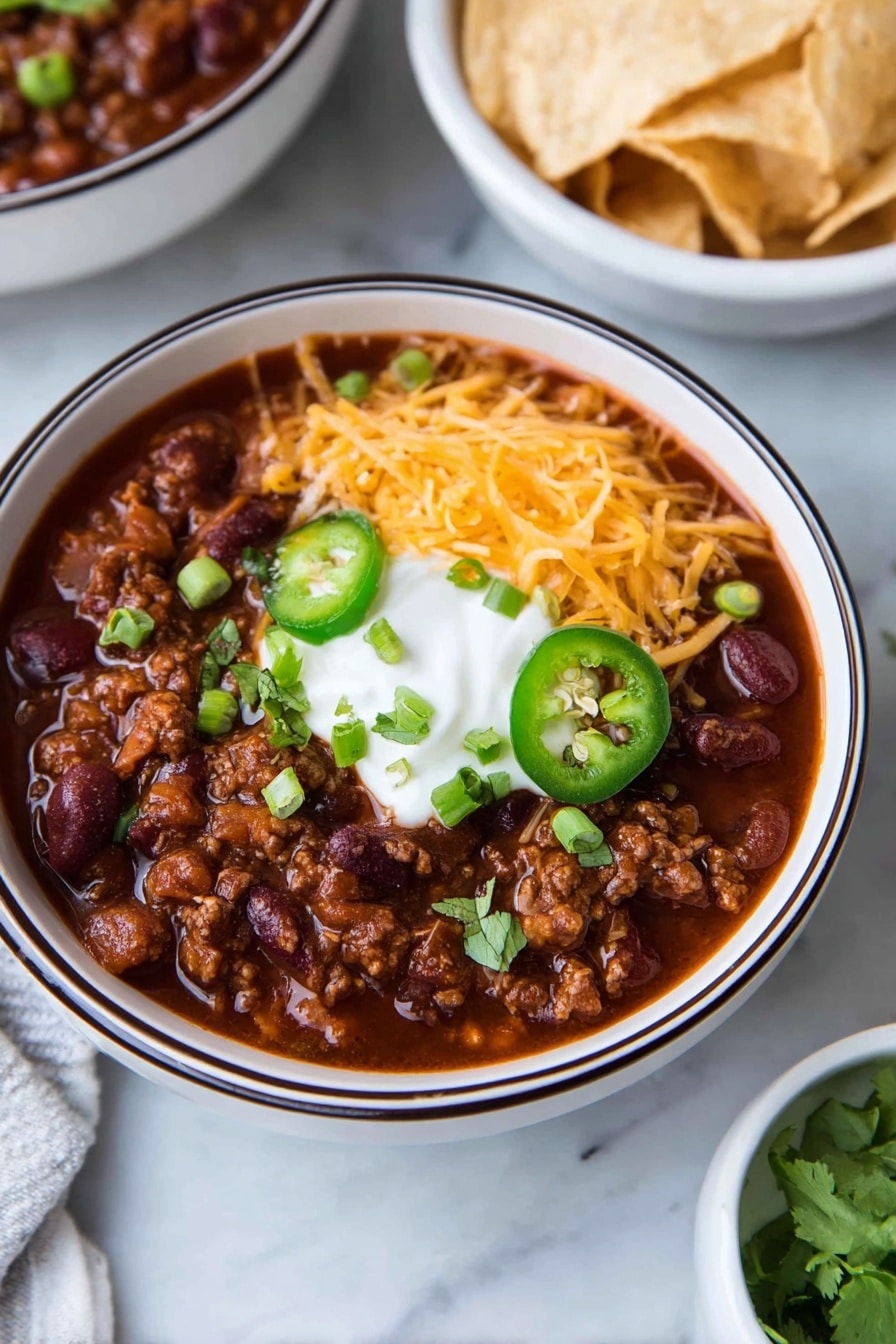 A white bowl with a dark rim holds a thick dark red chili filled with beans and ground meat. On top, there is a layer of shredded orange cheddar cheese on one side, a dollop of white sour cream in the center, sliced green jalapeño rings below, and chopped green onions and cilantro scattered around. In the background, there is a white bowl filled with light-colored tortilla chips on a white marbled surface. Photo taken with an iphone --ar 2:3 --v 7 - Healthy Ground Turkey Chili, ground turkey chili, healthy chili recipe, easy turkey chili, nutritious comfort food